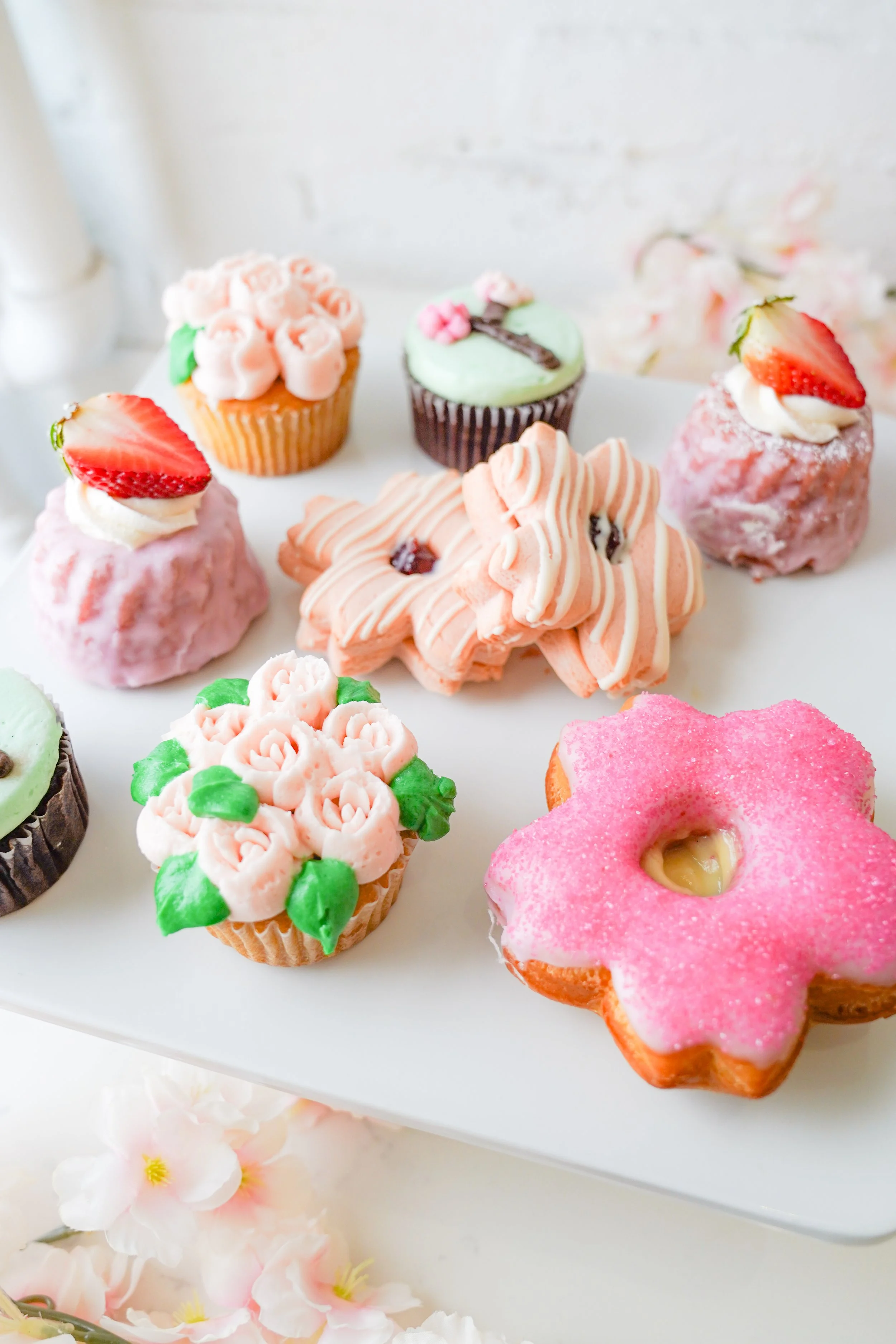 Assorted decorated cupcakes and cookies with pastel icing and strawberries, on a white tray.