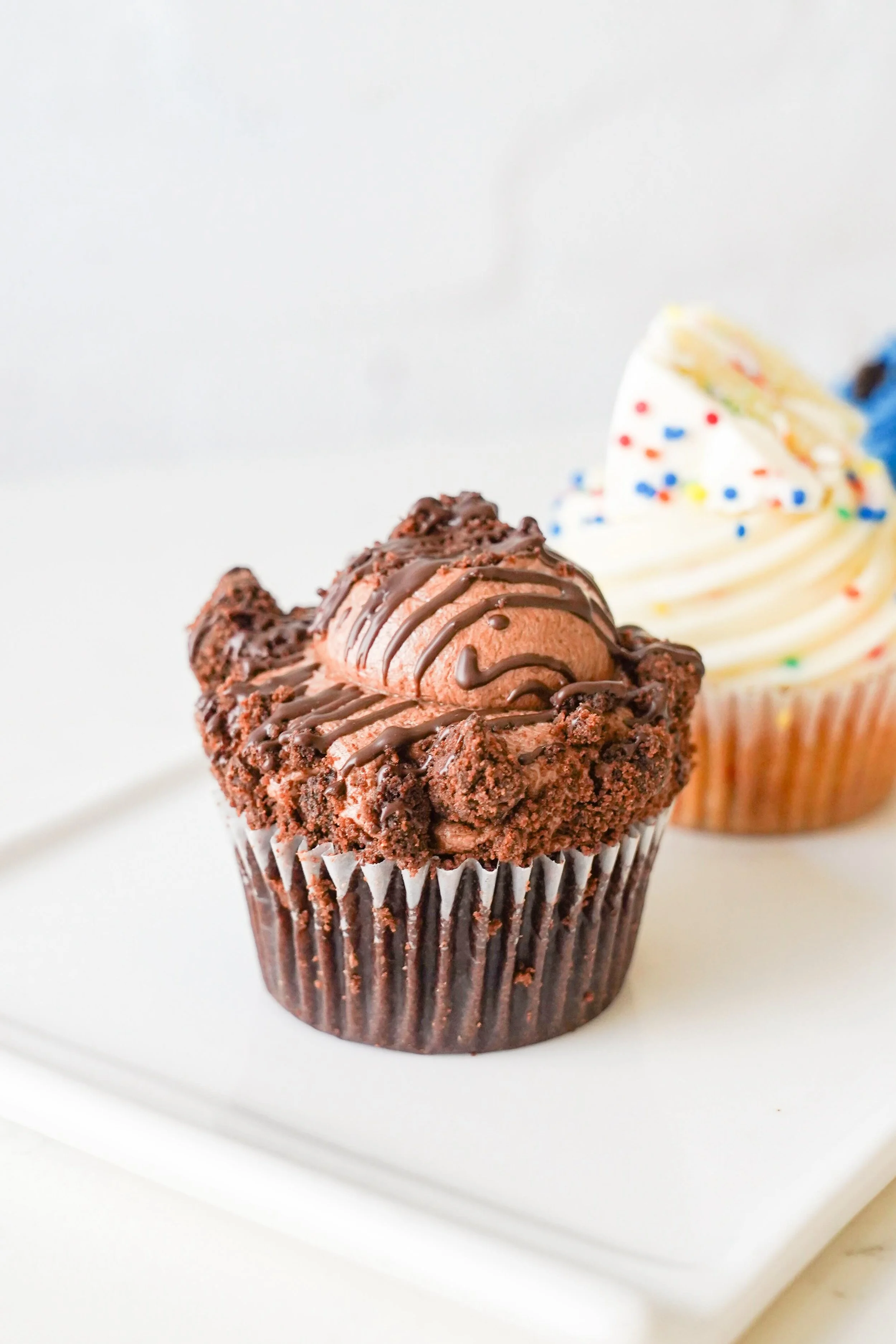 Chocolate cupcake with chocolate frosting and sprinkles on a plate, with a vanilla cupcake in the background.