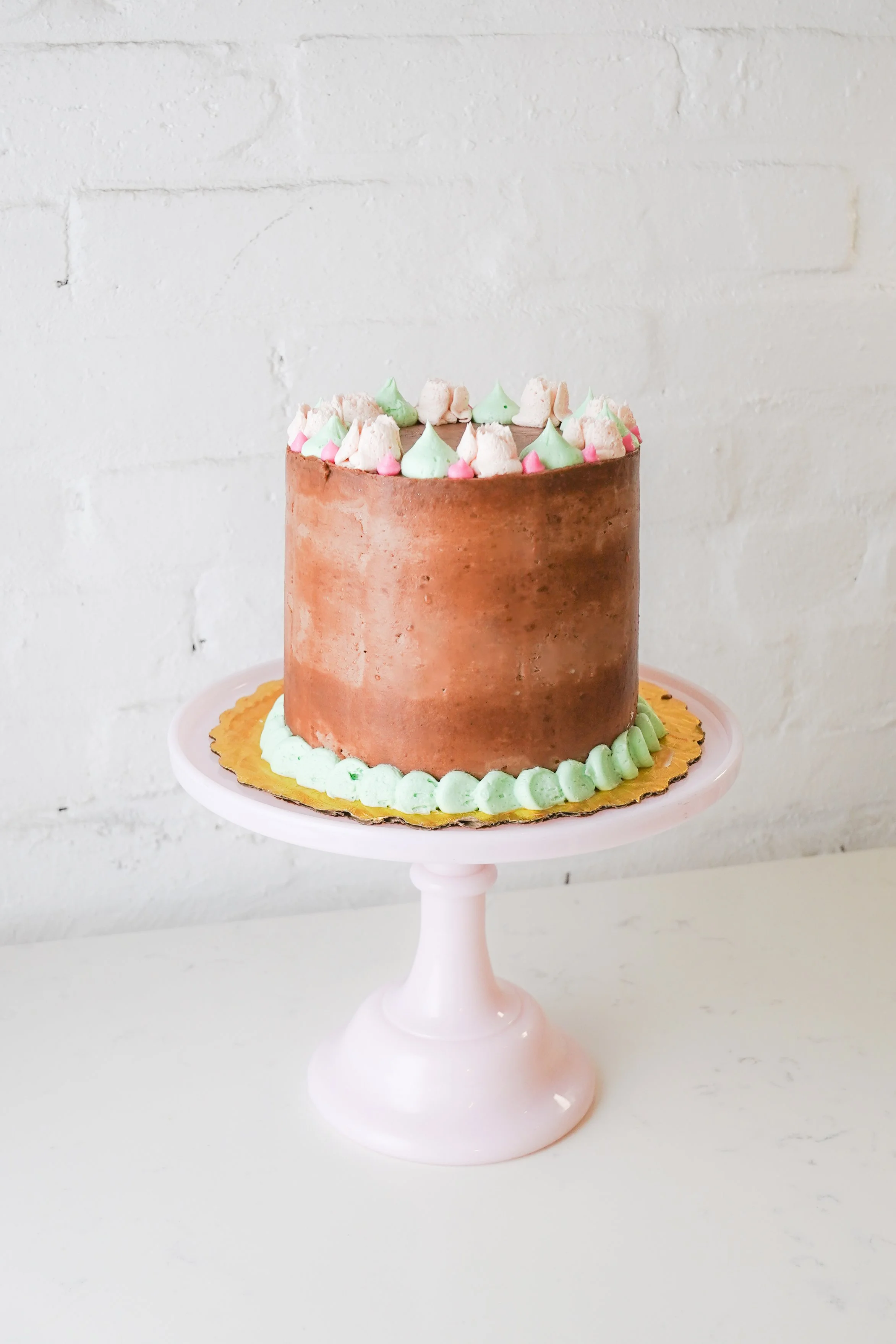 Chocolate cake with pastel frosting decorations on a pink cake stand against a white brick wall.
