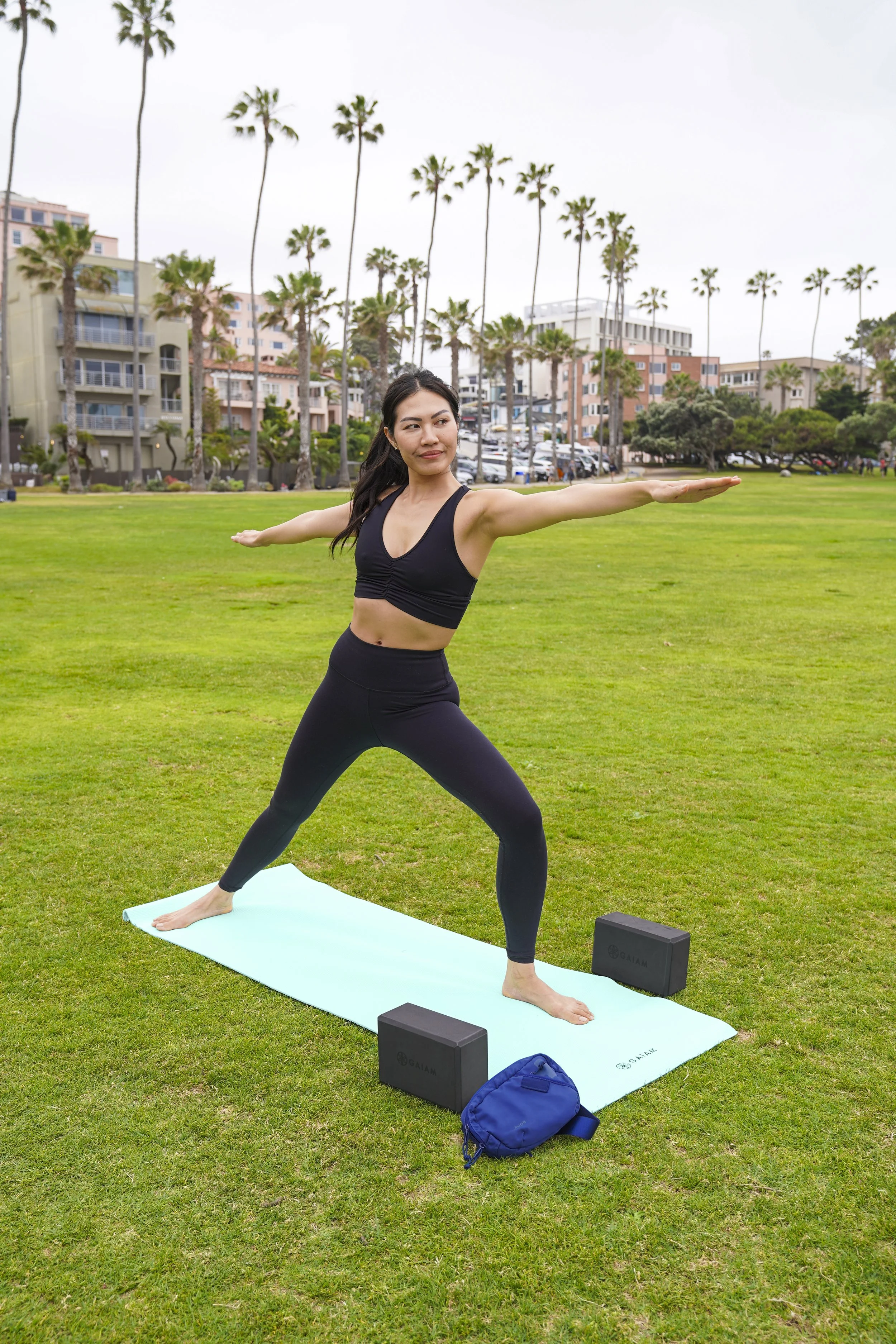 Person practicing yoga in a park, performing Warrior II pose on a yoga mat with blocks on grass, surrounded by palm trees and buildings in the background.