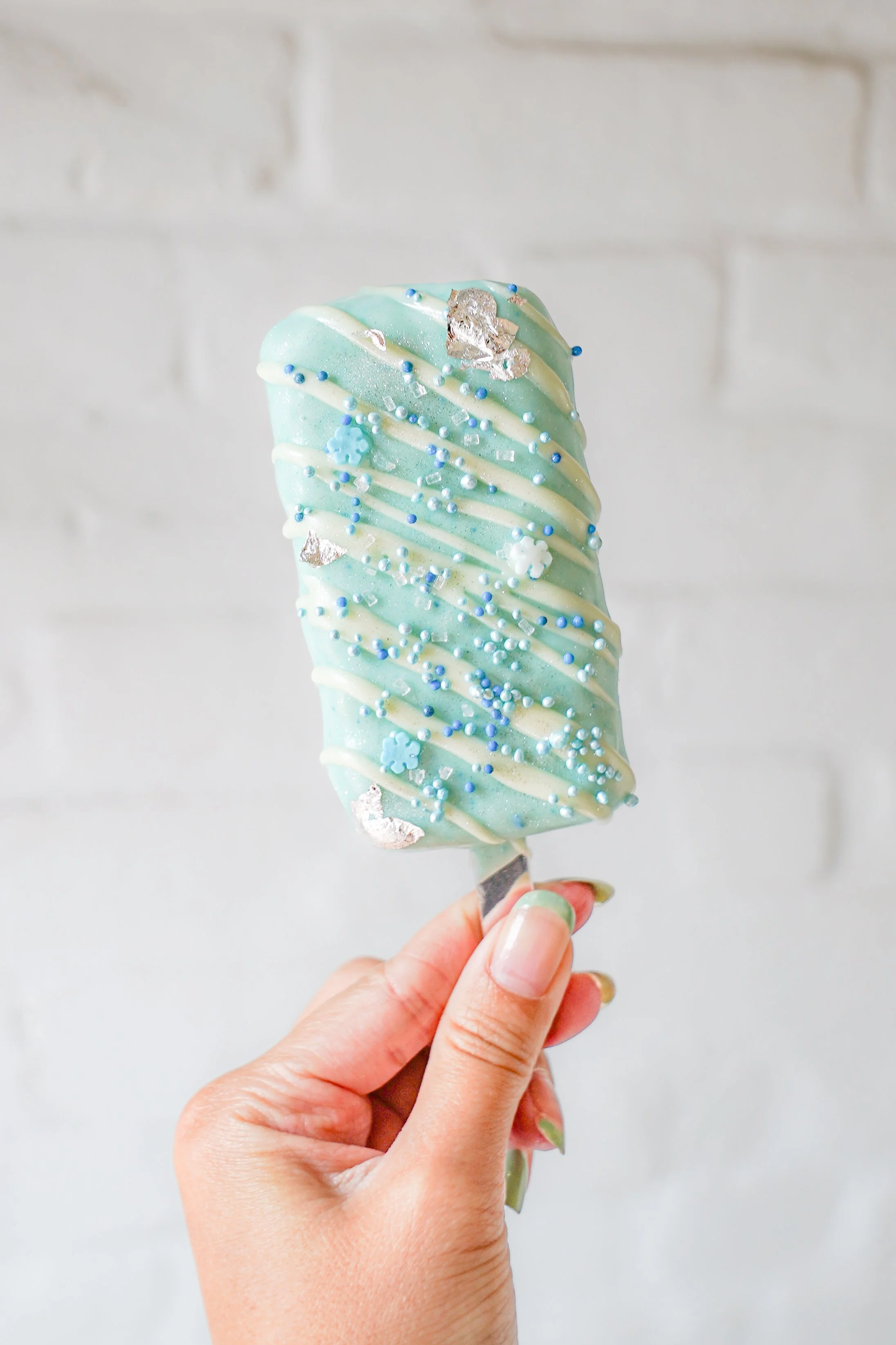 Hand holding an ice cream bar coated with light blue chocolate, white drizzle, and decorated with silver leaf, blue sprinkles, and flower-shaped sprinkles.