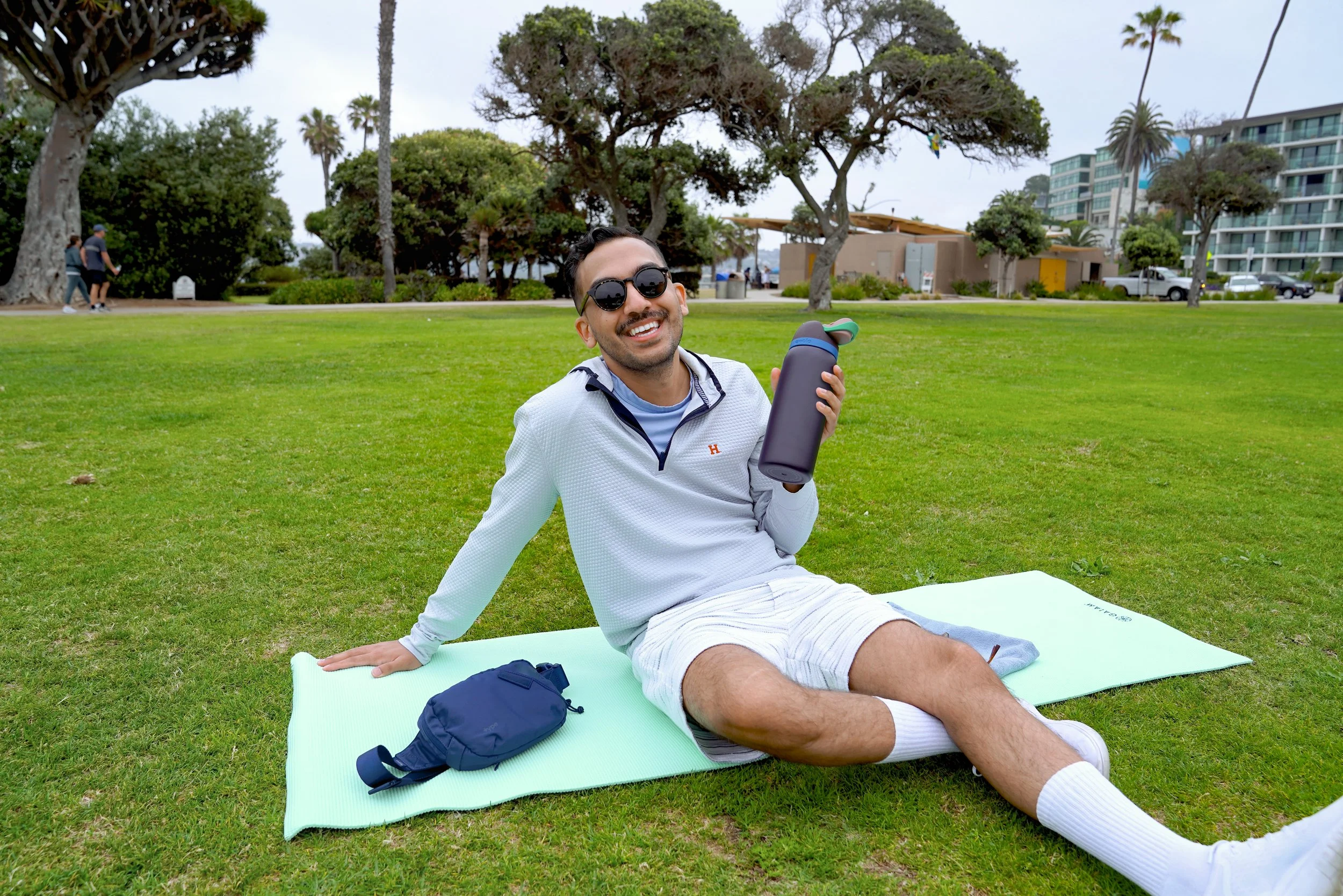 Man sitting on a mat in a park, smiling, holding a water bottle, wearing sunglasses and casual clothes.