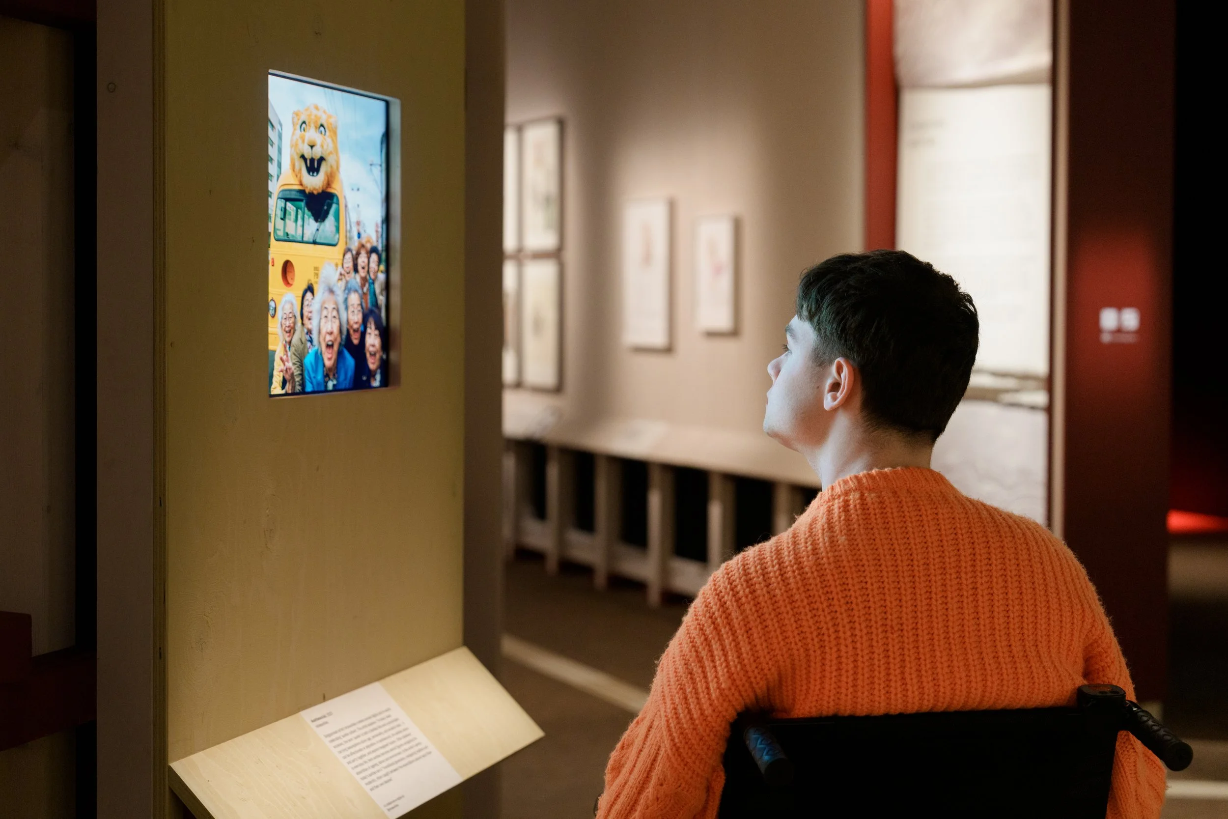 Young man in a wheelchair looking at a digital display of a photograph of elderly people and a lion mascot on a bus.