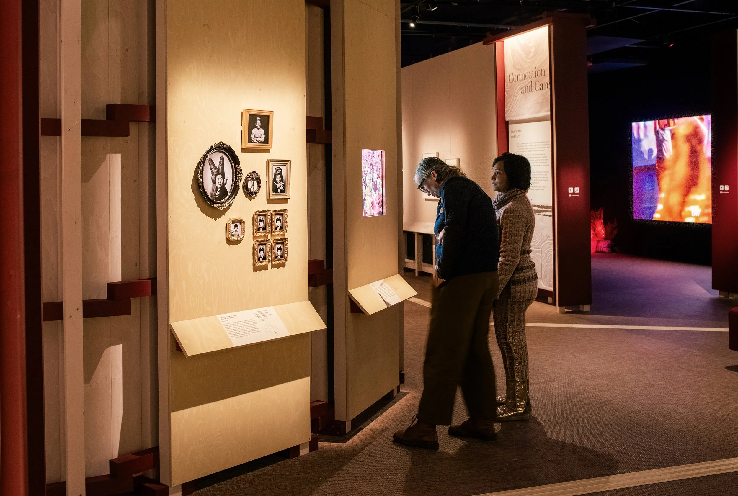 Two people observing framed photographs and information plaques in an art exhibit, with a screen displaying a colorful image in the background.