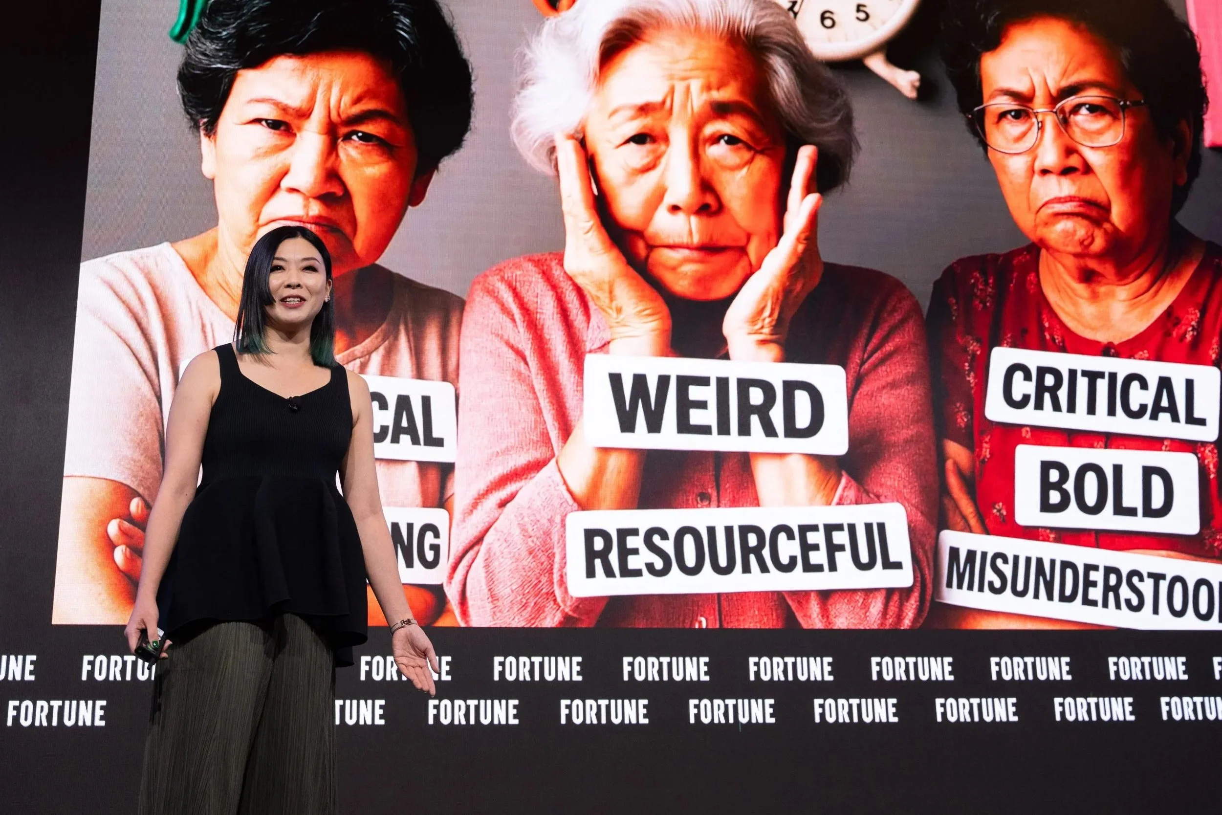 A woman standing on stage in front of a large screen displaying three elderly women with labels across their chests. The labels read 'FUNNY,' 'WEIRD RESOURCEFUL,' and 'CRITICAL BOLD MISUNDERSTOOD.' The stage backdrop has the word 'FORTUNE' repeatedly printed on it.