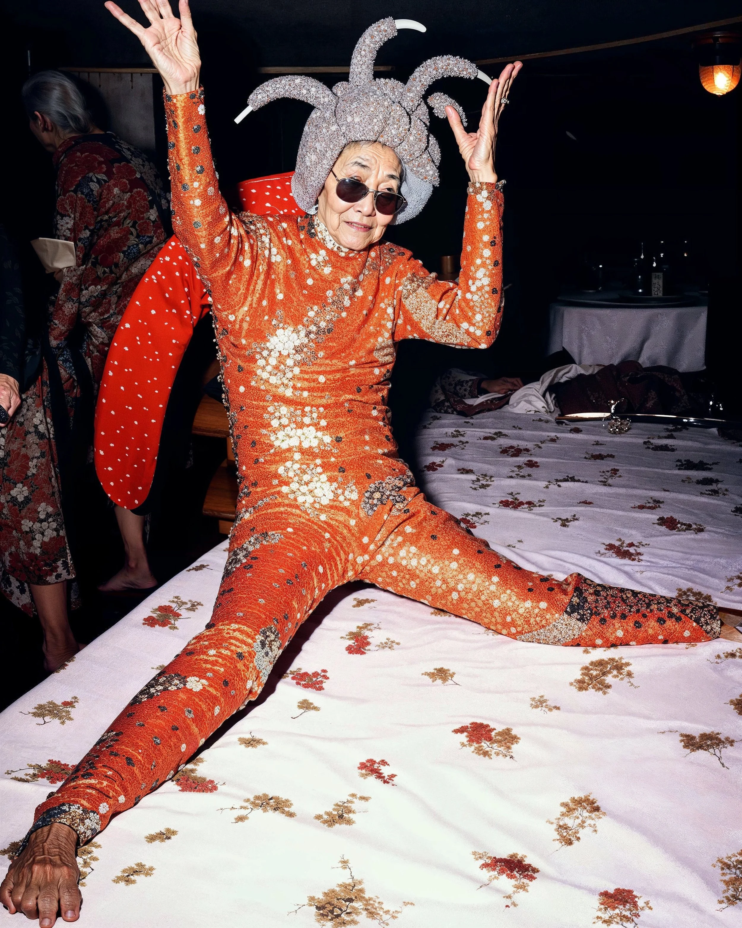 An elderly woman dressed in an orange, sparkly jumpsuit with a matching bejeweled headpiece and sunglasses, sitting on a bed with her legs spread wide and her arms raised in a joyful dance pose.