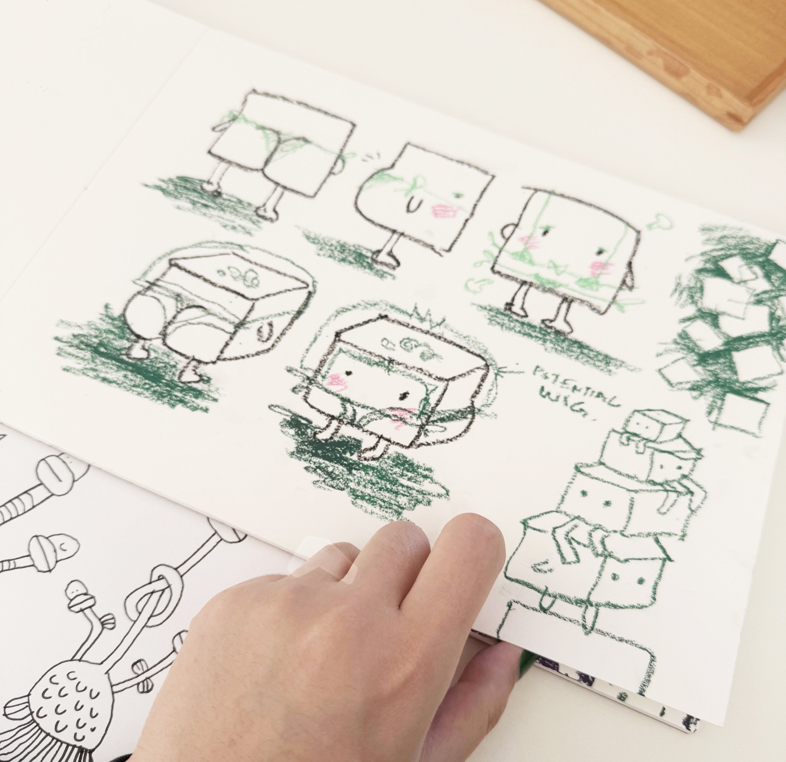 Creative hand-drawn illustrations of various square-shaped characters with different facial expressions, standing, sitting, and with a belly button, drawn in green and pink crayons on a white sheet of paper.