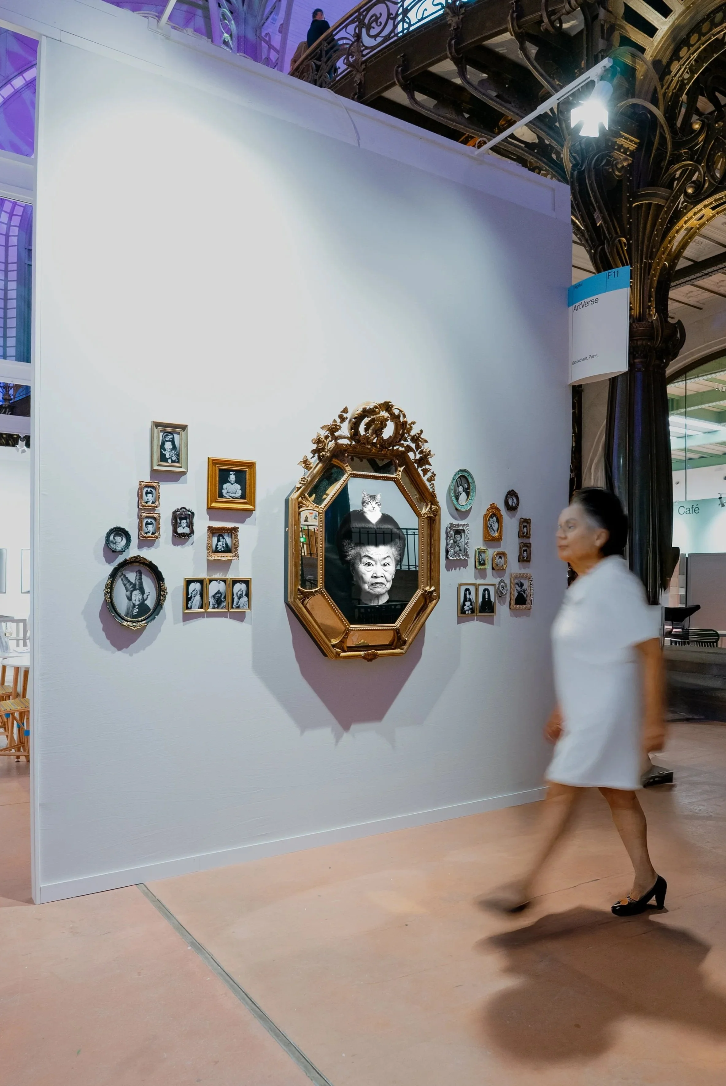 An art display featuring a large mirror with an ornate golden frame. Around the mirror are various black-and-white family photographs in different sizes and frames on a white wall. A woman in a white dress and black heels is walking past the display.