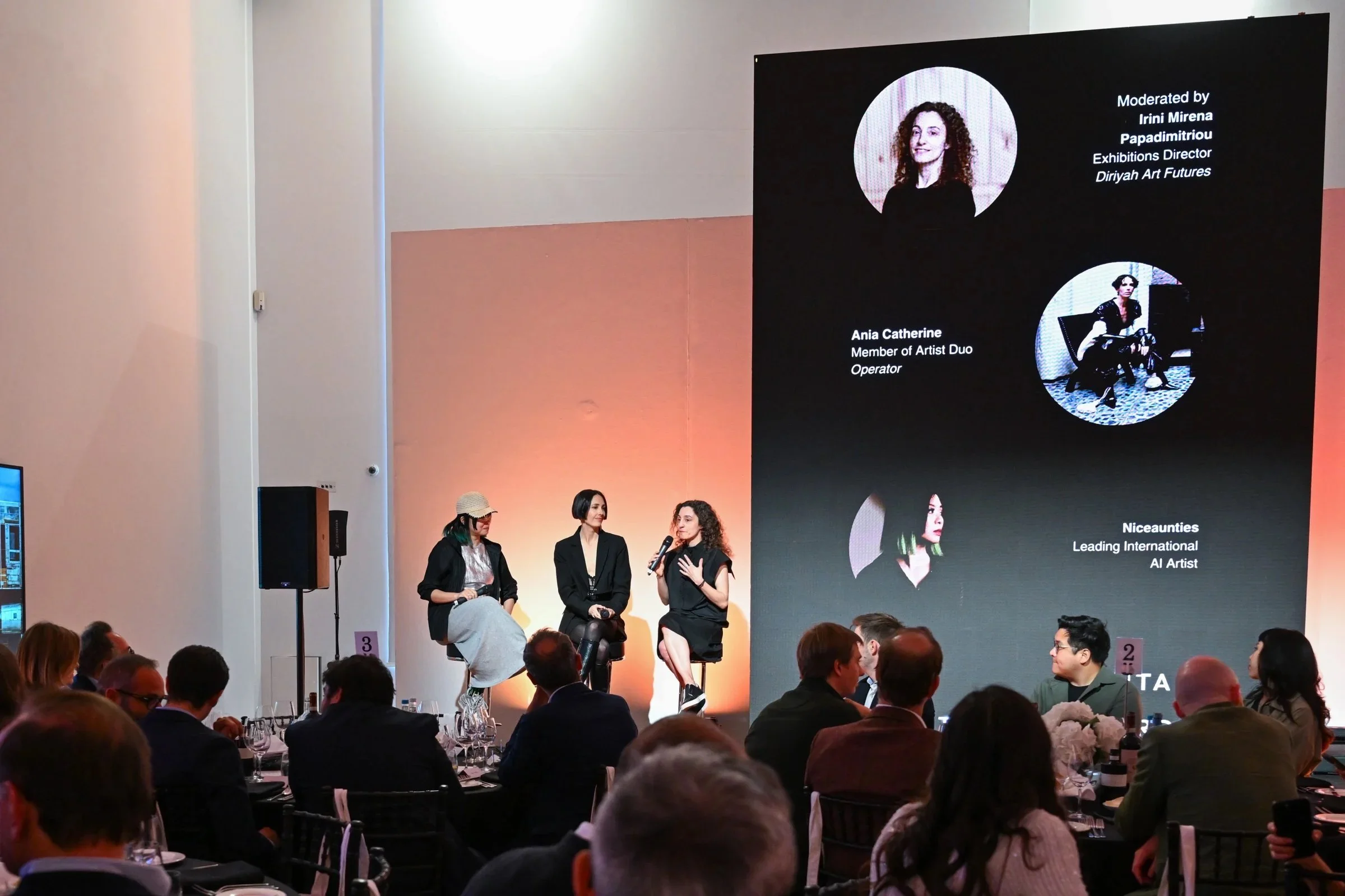A panel discussion with three women speaking on stage in front of an audience. One woman is holding a microphone, and a large screen behind displays details of the panelists, including their names and roles.