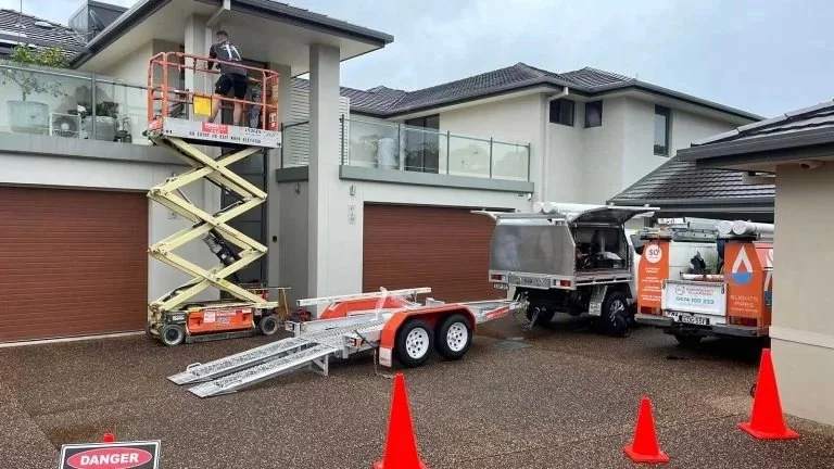 A worker on a scissor lift cleaning or inspecting a glass railing on a modern house's balcony, with a service truck and trailers parked outside on the driveway, and orange cones marking the area.