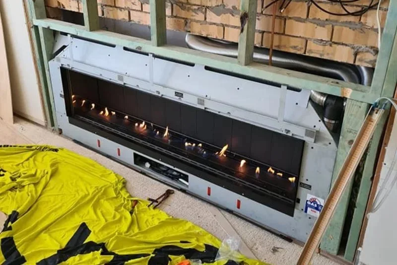 Installation view of a fireplace insert with visible flames, installed in a framed wall with wooden studs, brick wall background, and construction tools nearby.