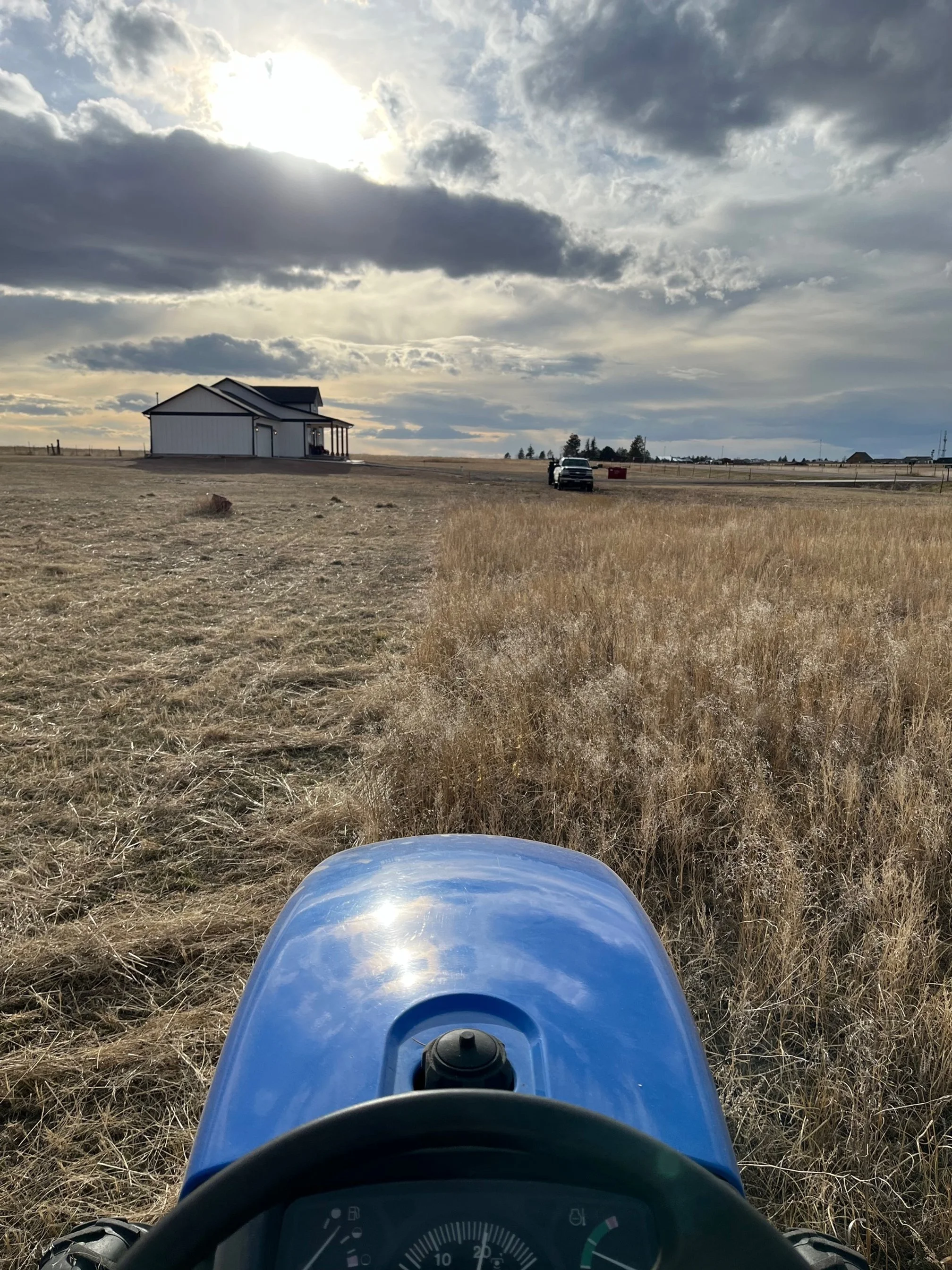 View from a tractor in a field showing a house in the distance, a truck, a red container, and cloudy sky with the sun partially obscured.
