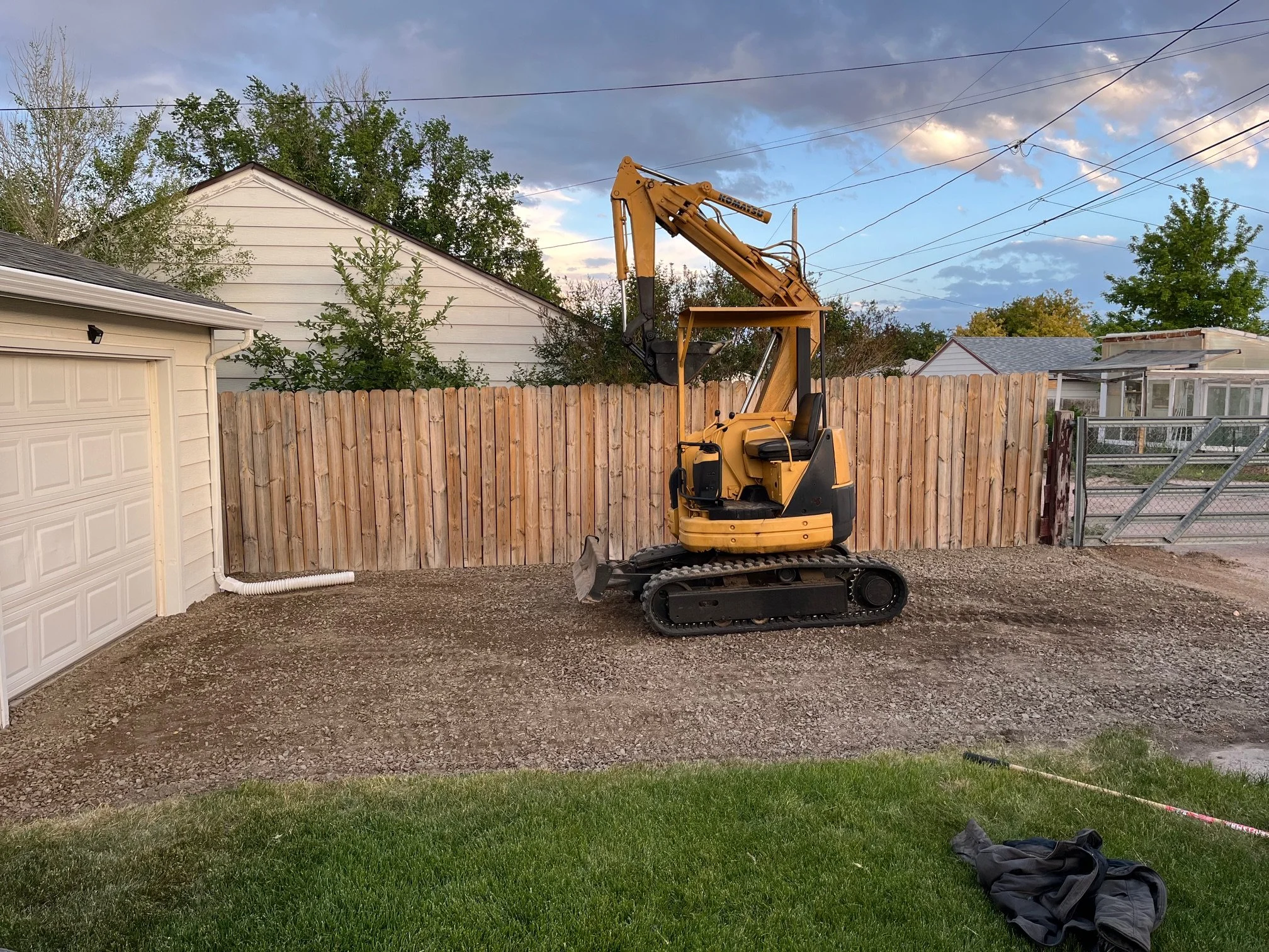 A small yellow excavator parked on a gravel surface in a backyard, with a wooden fence, trees, and houses in the background.