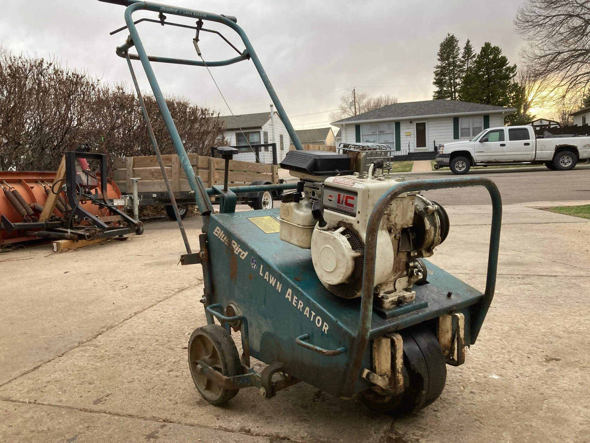 A small blue gas-powered lawn aerator machine on a concrete driveway, with a white house, trees, a white pickup truck, and other equipment in the background.