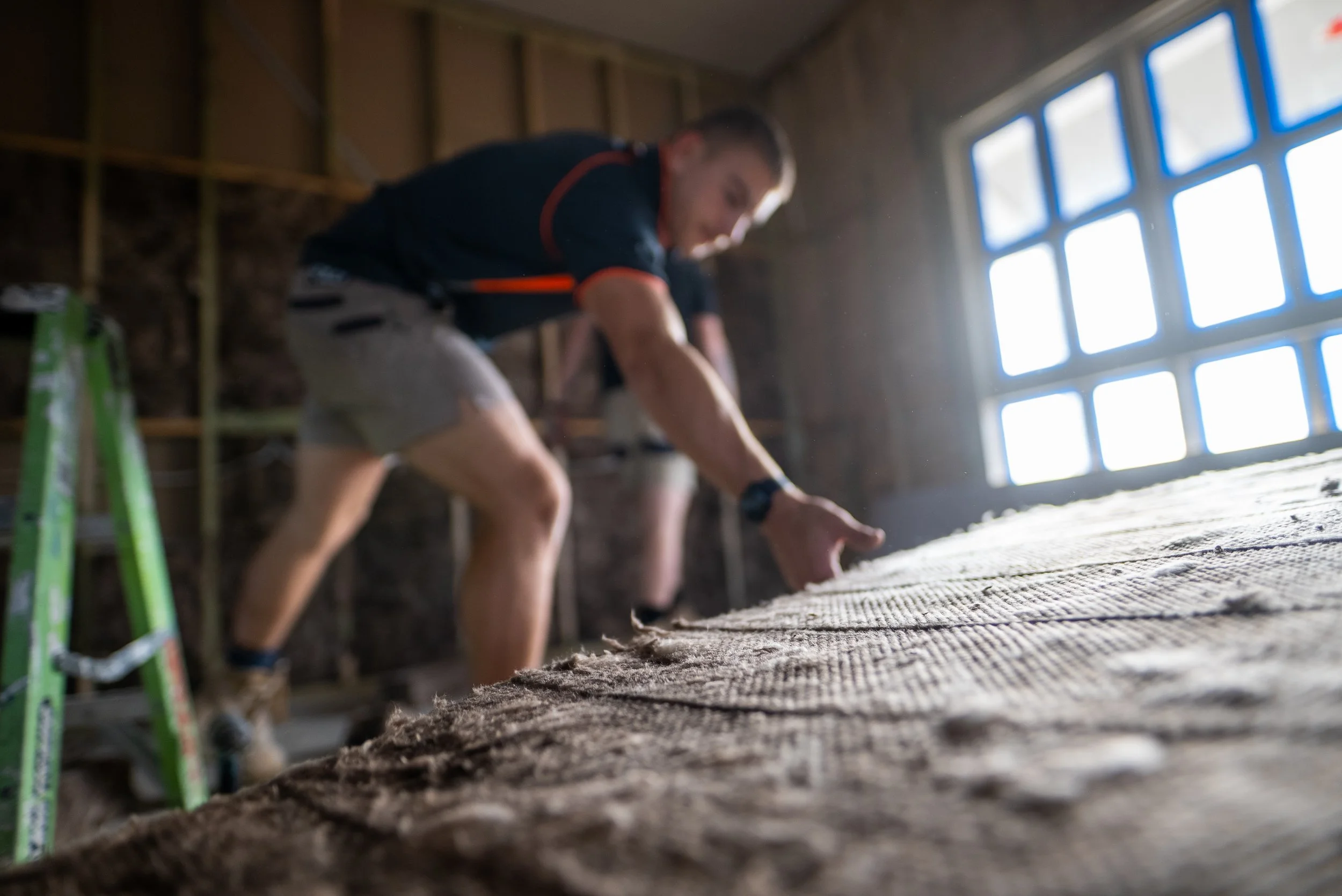 Man working on a construction project inside a wooden structure, examining a large piece of fabric or insulation on the floor, with natural light coming through a window.