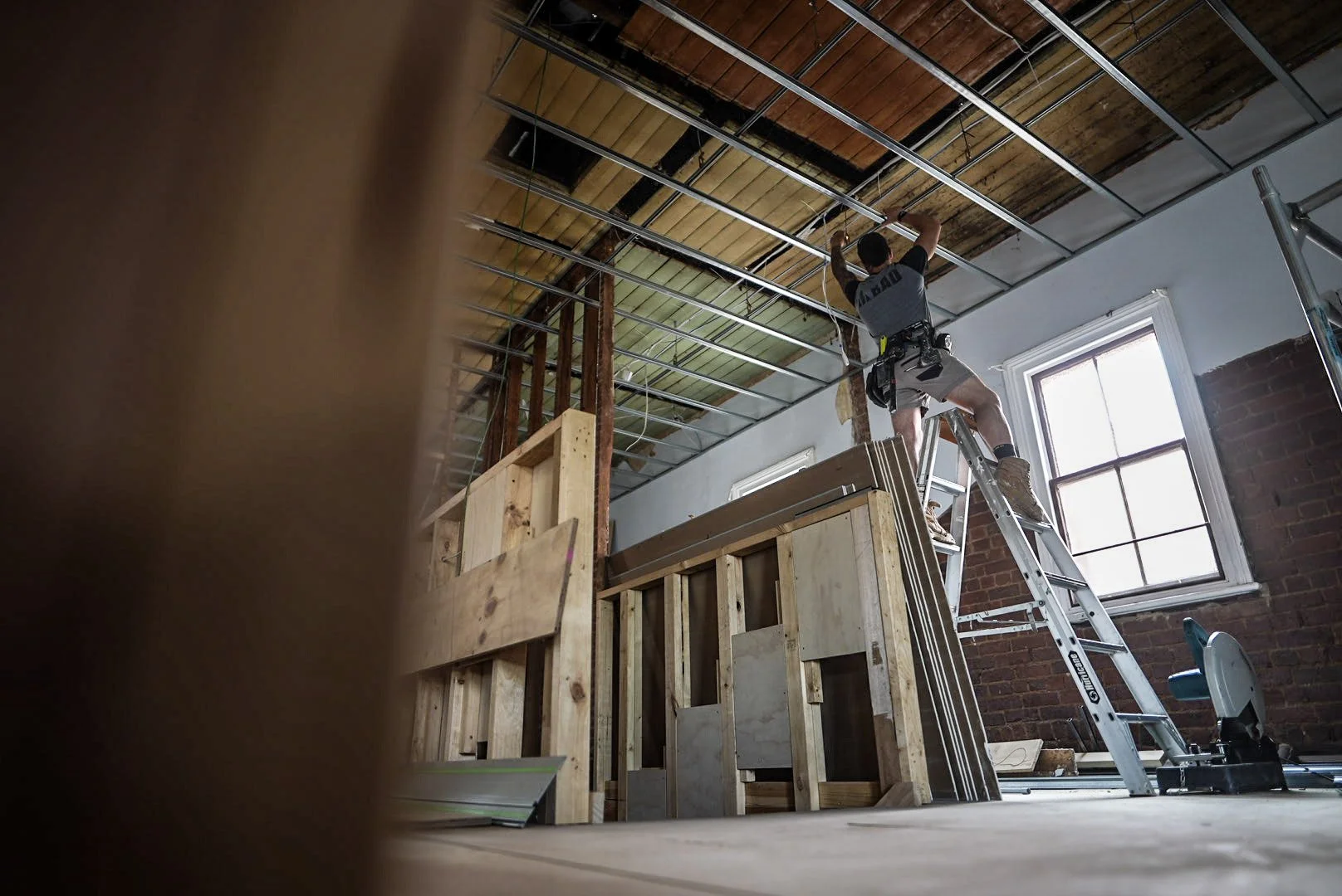 A construction worker standing on a ladder installing ceiling framing in a room under renovation with exposed brick walls and wooden flooring.
