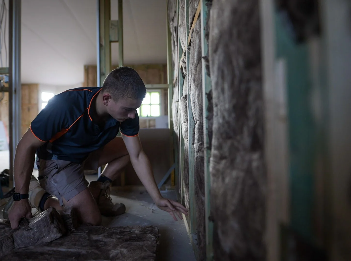 A man working on a construction site, installing insulation within a wall frame inside a building under construction.