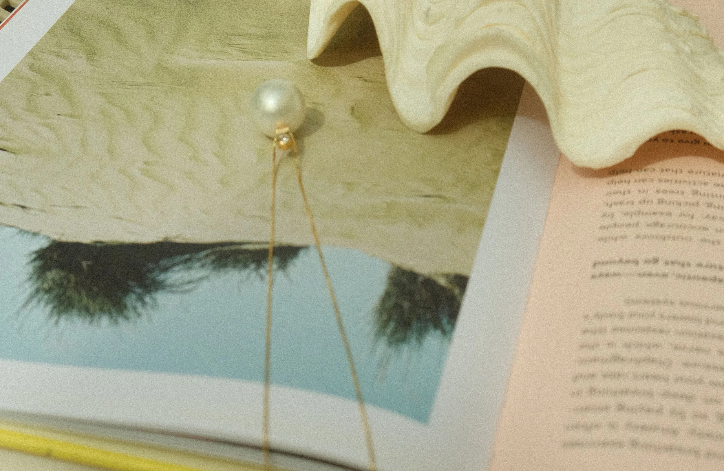 Close-up of a photograph with a pearl and gold chain, a piece of wood with a wavy edge, and a partially visible soft pink paper with blue sky, trees, and some handwritten or printed text.