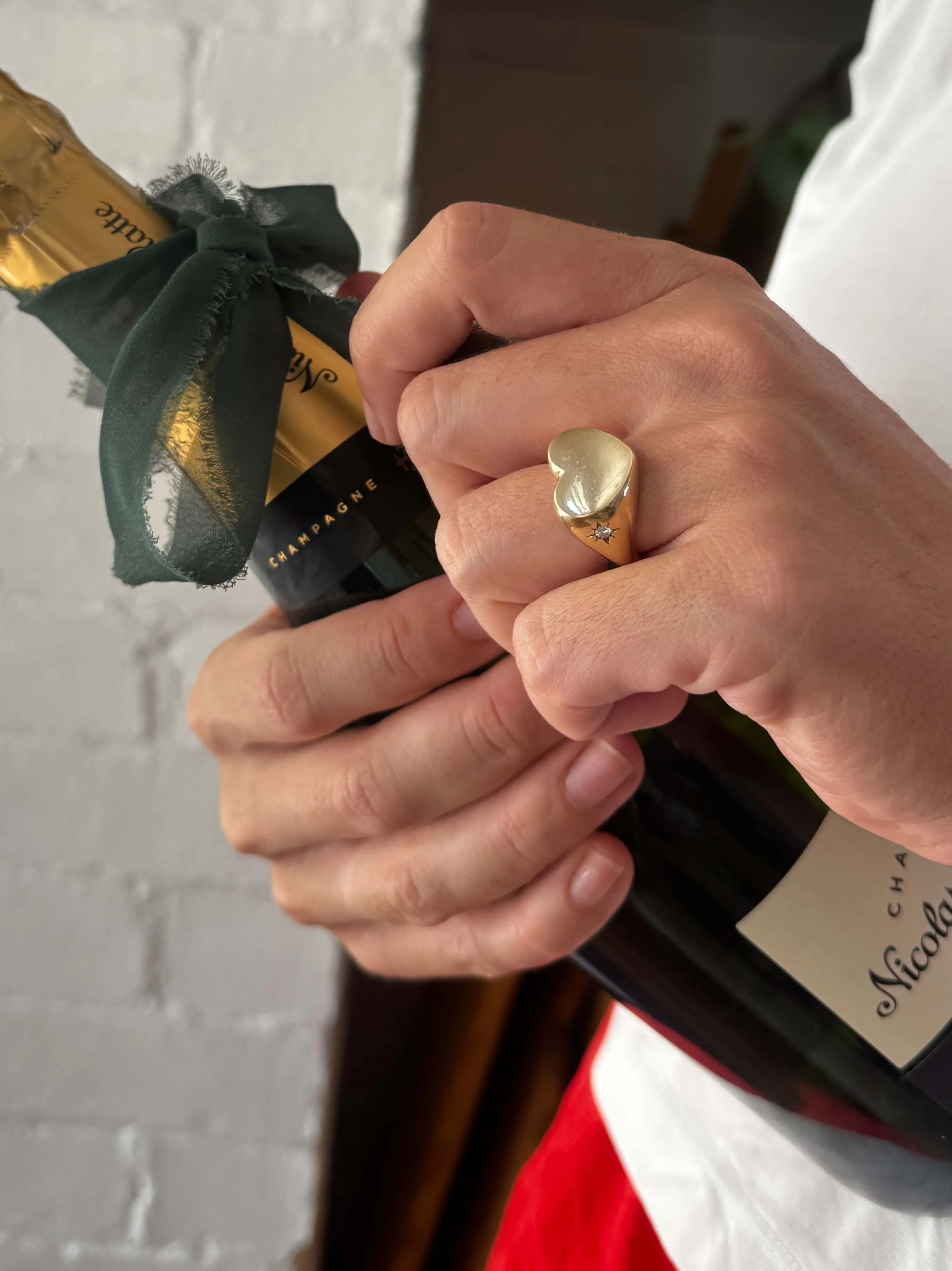 Person holding a bottle of Nicolas Feuillatte champagne with a gold cap and a green ribbon, wearing a gold ring with a heart shape and a small star-shaped detail.