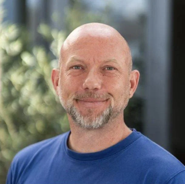 A bald man with a beard and mustache smiling outdoors, wearing a blue shirt.