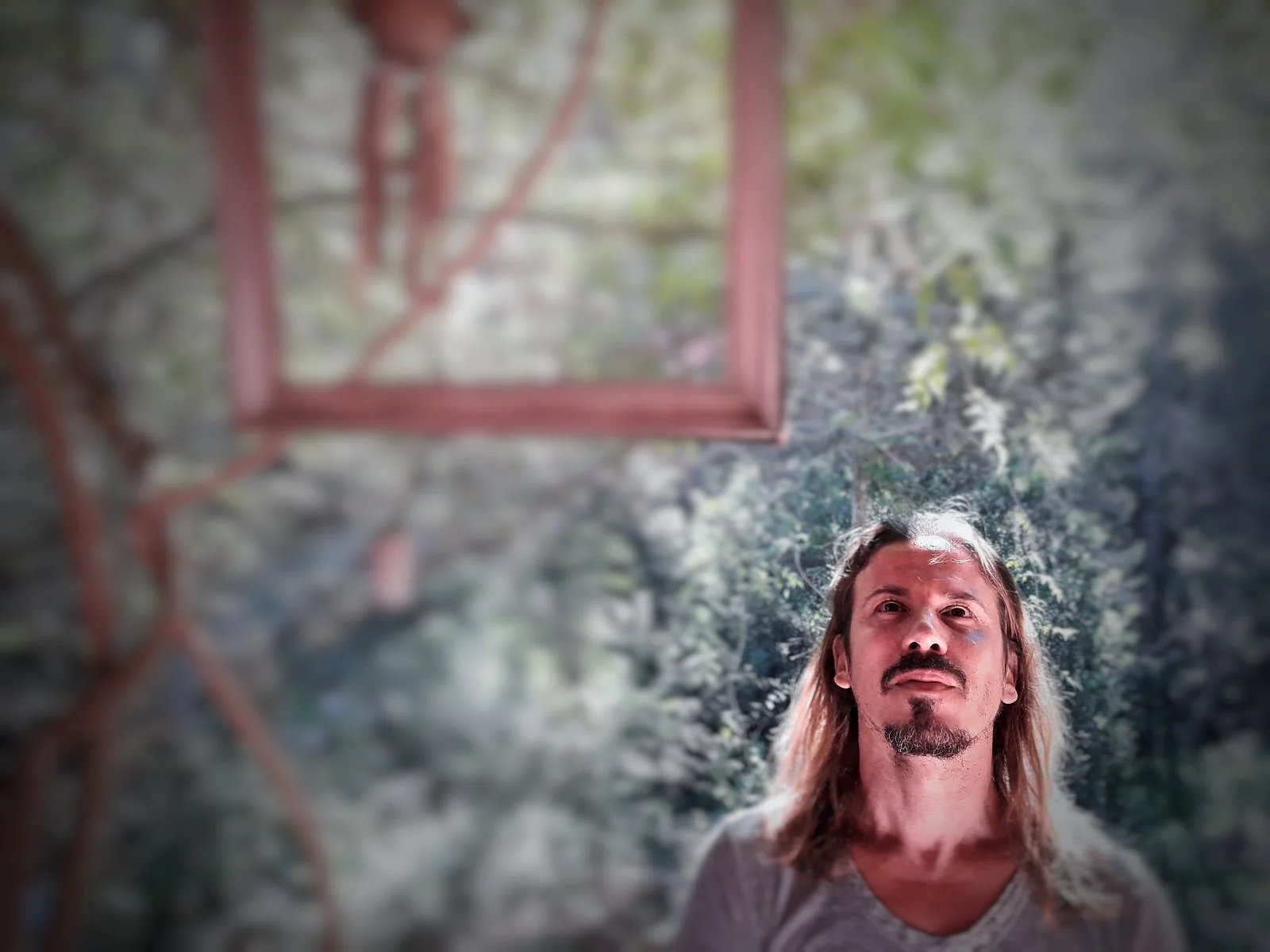 A man with long, blonde hair, a goatee, and mustache, wearing a light-colored shirt, looking directly at the camera with a serious expression. The background is outdoors with green foliage and a structure with red wooden frames are visible, out of focus.