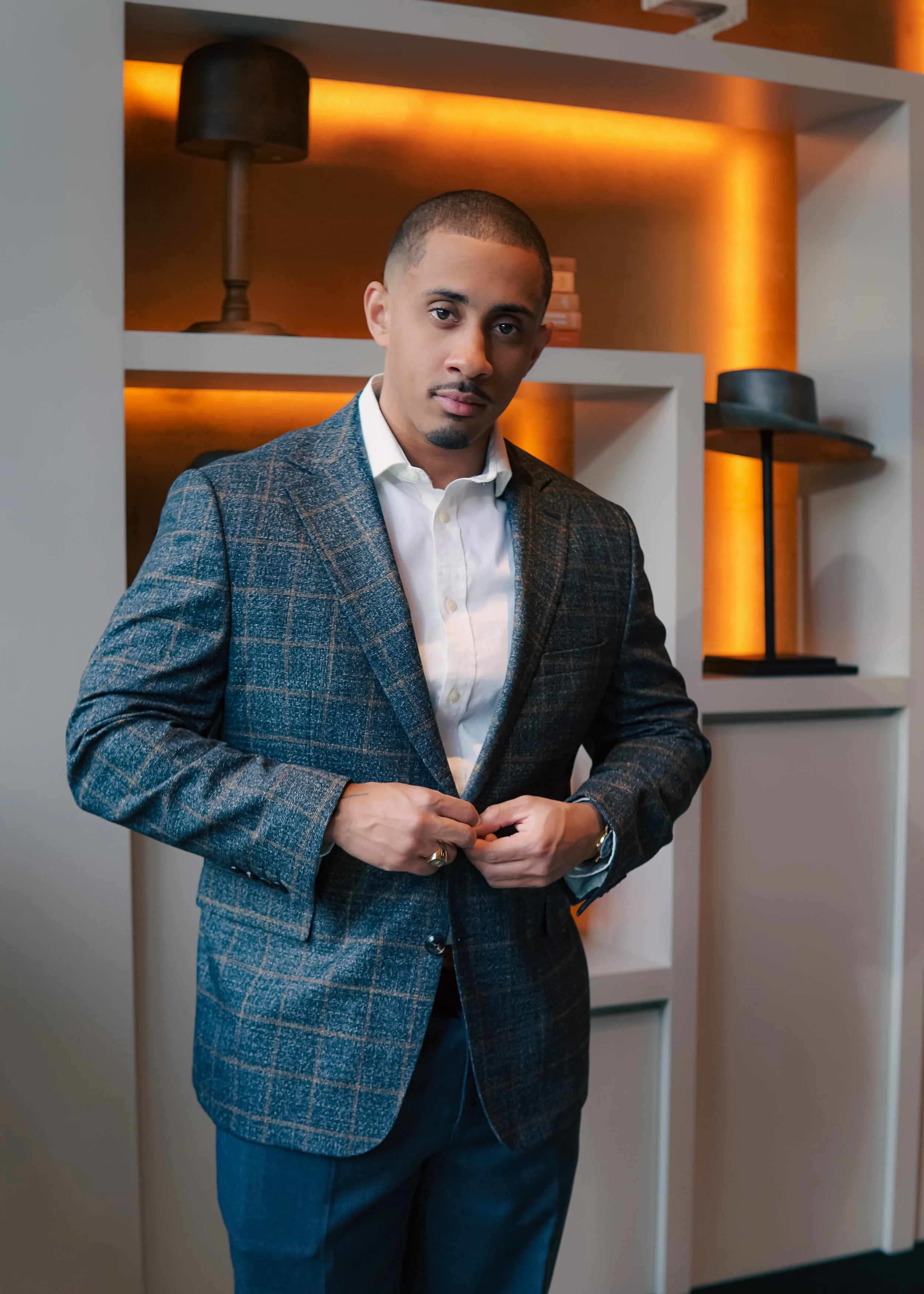 A man in a plaid suit buttoning his jacket in a stylish indoor setting with warm lighting and shelves in the background.