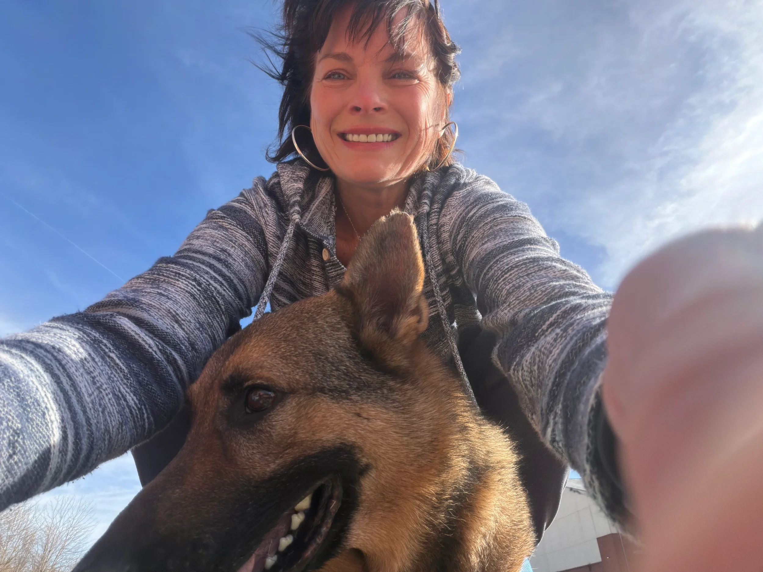An FVC client smiling and taking a selfie outdoors with her dog, a German Shepherd, against a blue sky with wispy clouds.