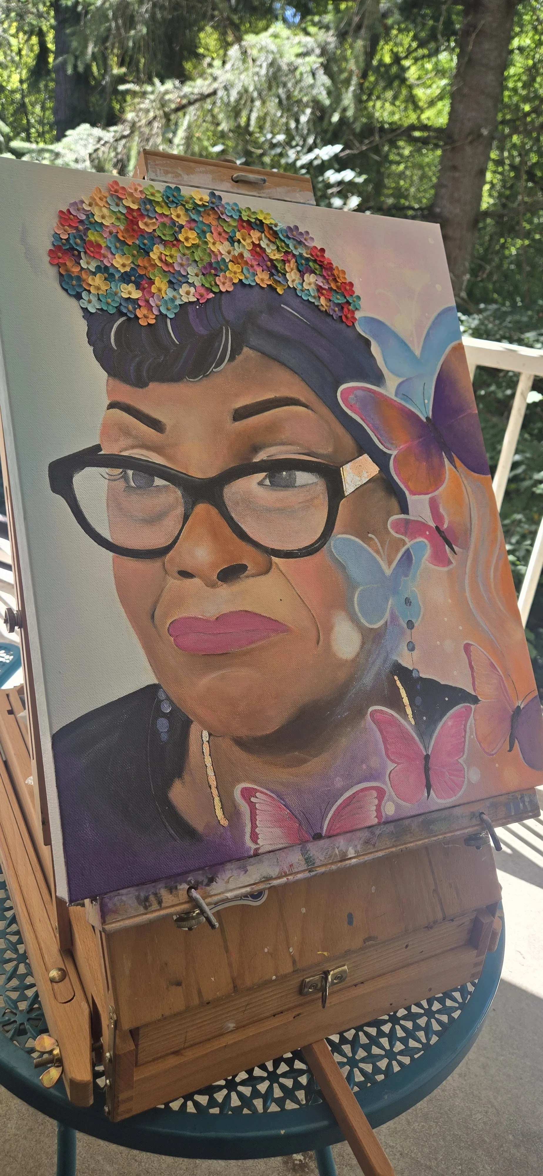 Portrait painting of my mother, with glasses, colorful flower headdress, and butterflies, placed on an easel outdoors.