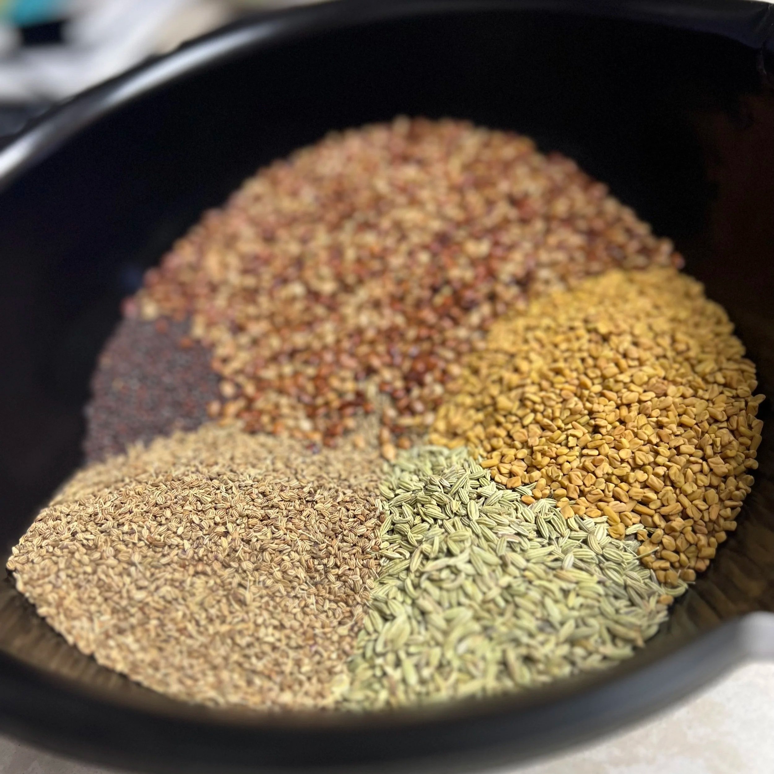 Close-up of a spice or seed mixture in a black bowl, with various grains and seeds in shades of brown, yellow, and green.