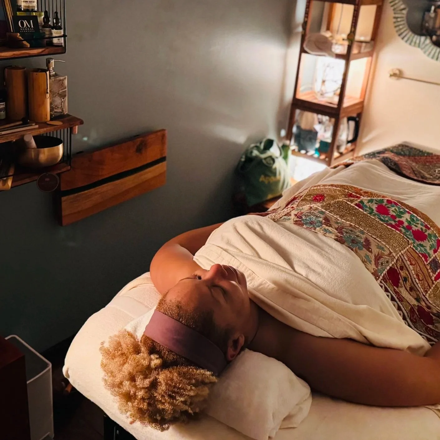 A person with curly blonde hair and a headband is lying in bed with eyes closed, resting on a pillow. They are covered with a patterned blanket and appear to be relaxed or sleeping. The room has shelves with bottles and containers, a green bag on the floor, and a wooden shelf unit with various items. The lighting is warm and cozy.