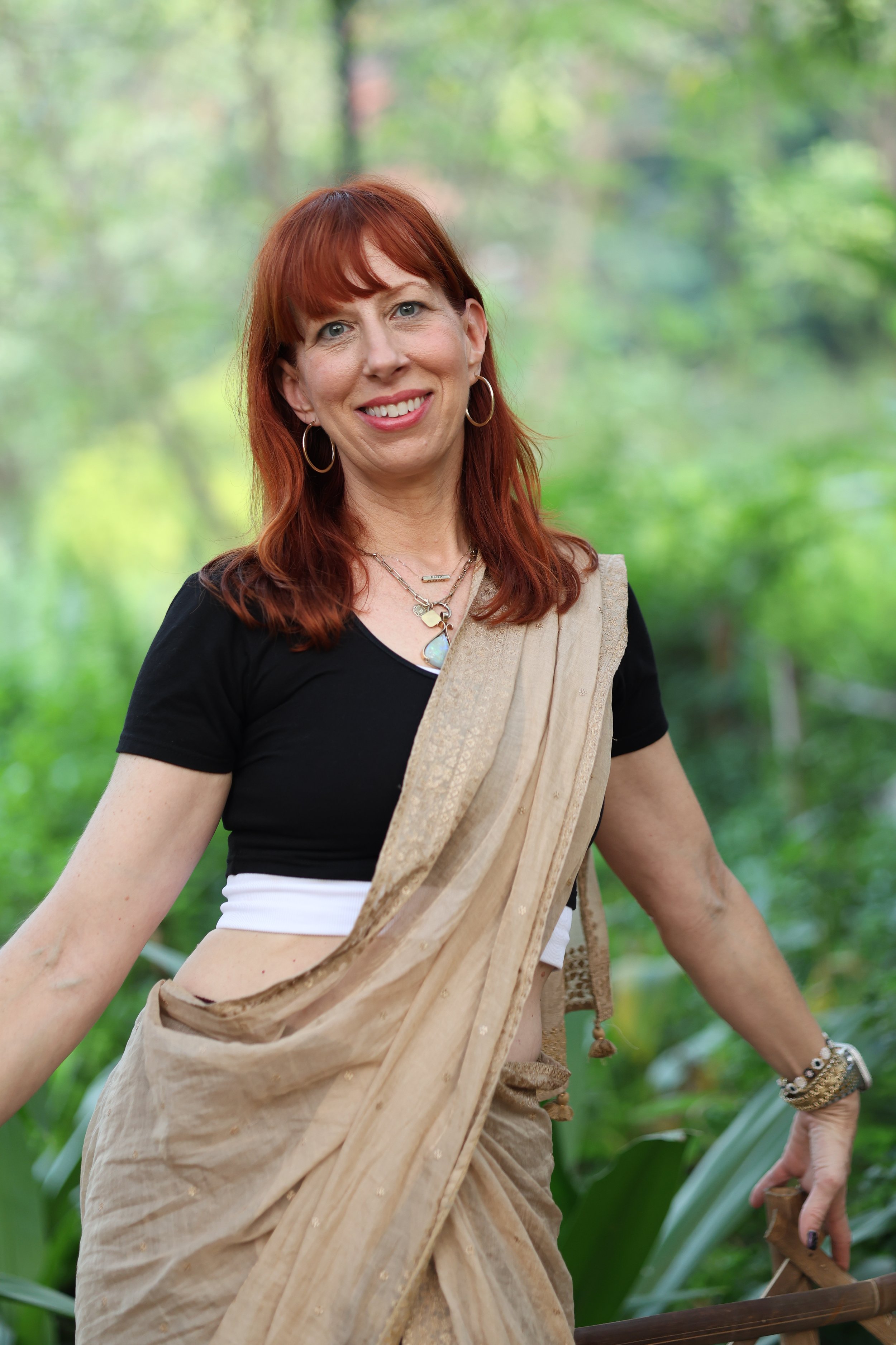 A woman with red hair, wearing hoop earrings, necklaces, a black crop top, and a beige sari, standing outdoors with green foliage in the background.