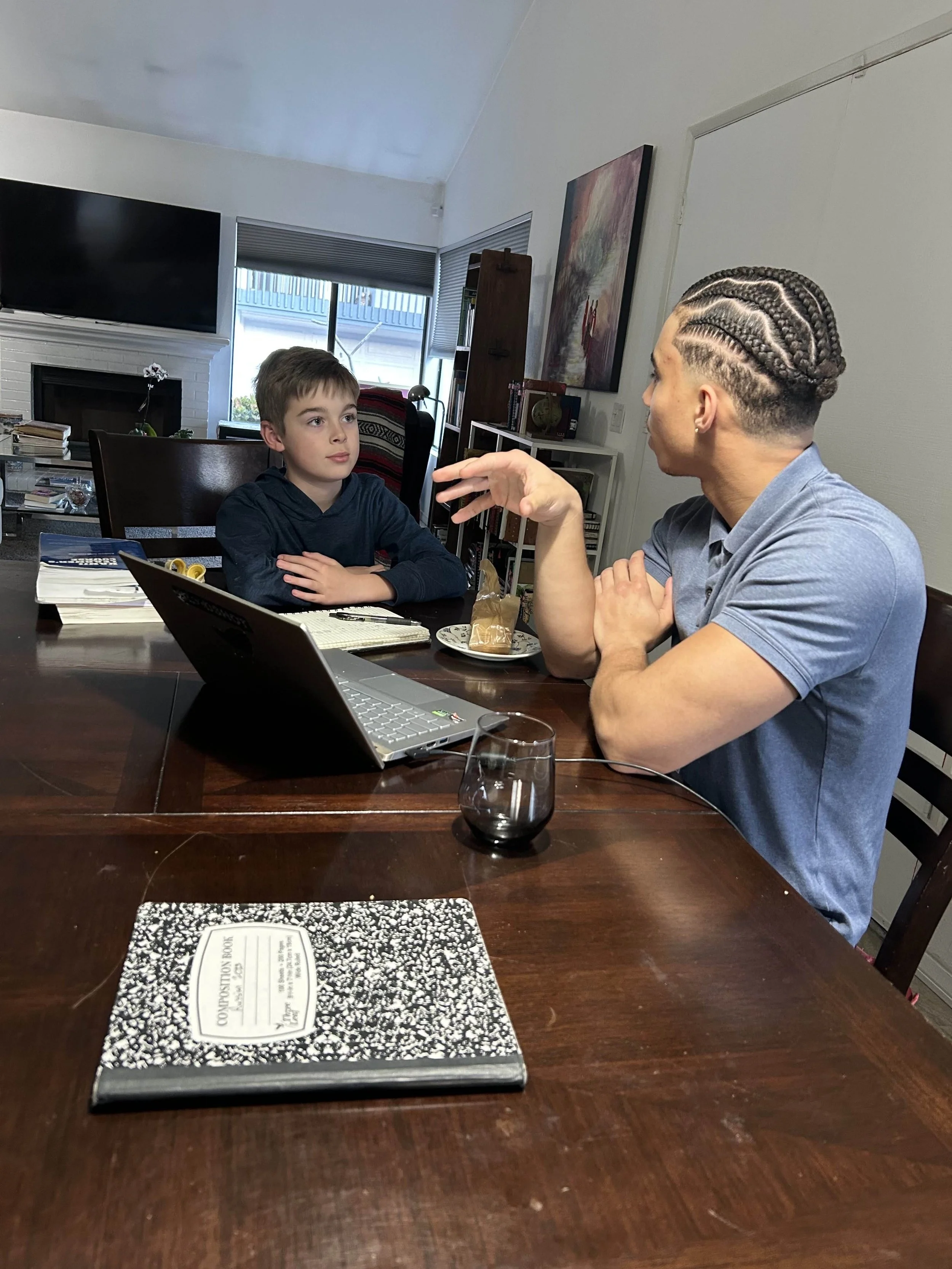 Two people sitting at a table with a laptop, notebook, and drink, studying Japanese in conversation in a home setting.