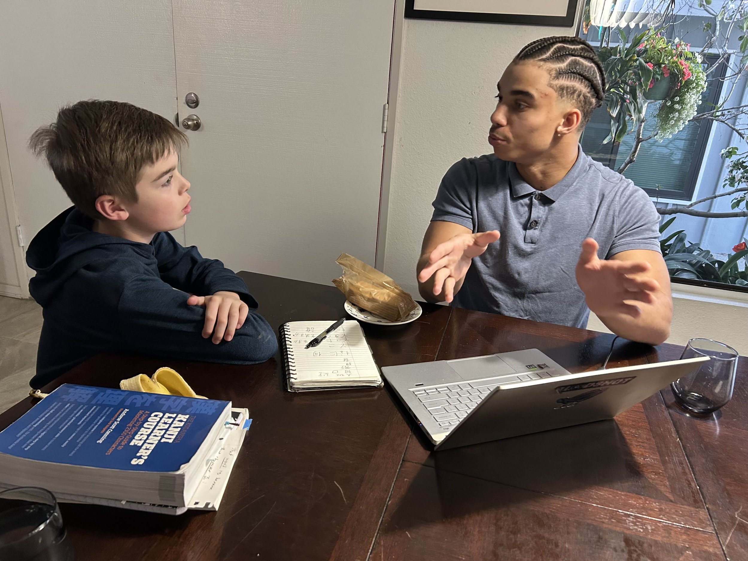 A young adult and a child engaged in a discussion at a table with a laptop, a textbook, and a notebook. The adult is speaking, gesturing with hands, while the child listens attentively. They are studying Japanese.