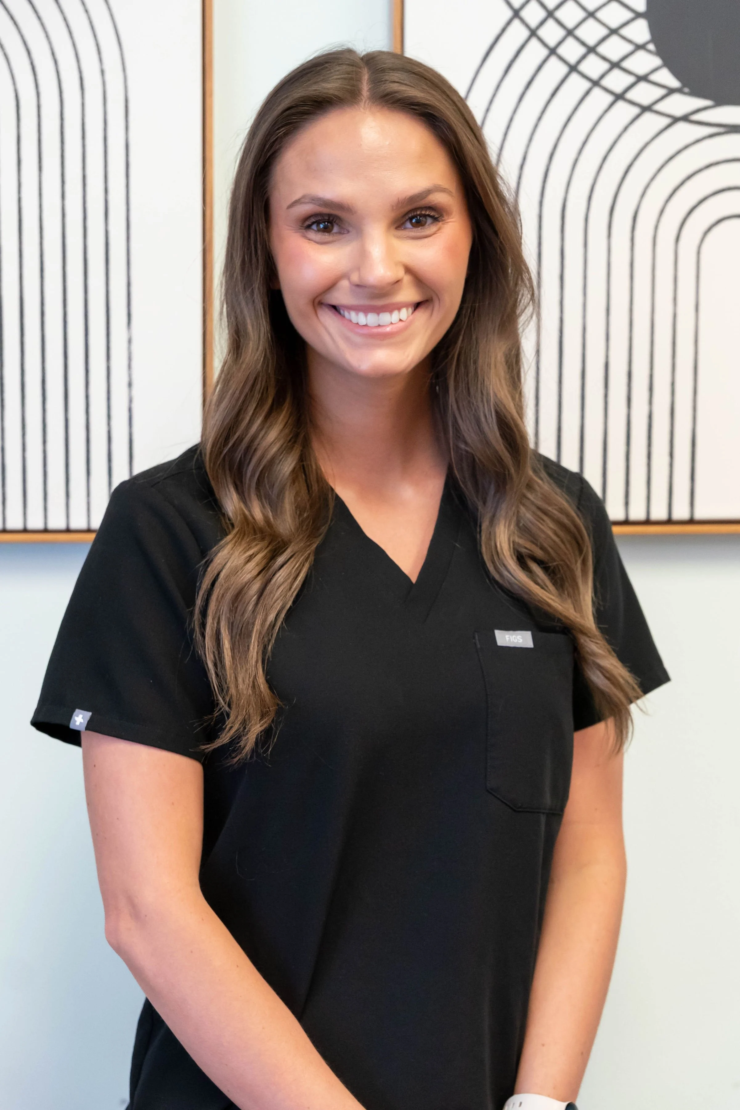 A smiling woman in a black medical scrub top standing in front of an abstract art background.