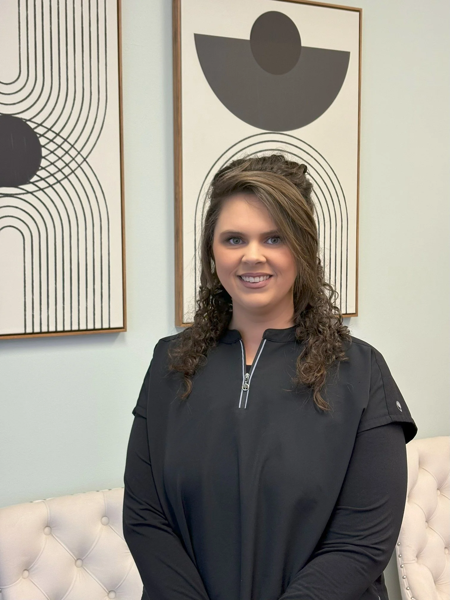 A woman with curly brown hair and a black zip-up top sitting on a white tufted couch in front of two abstract art pieces with black, white, and gray geometric designs on the wall.