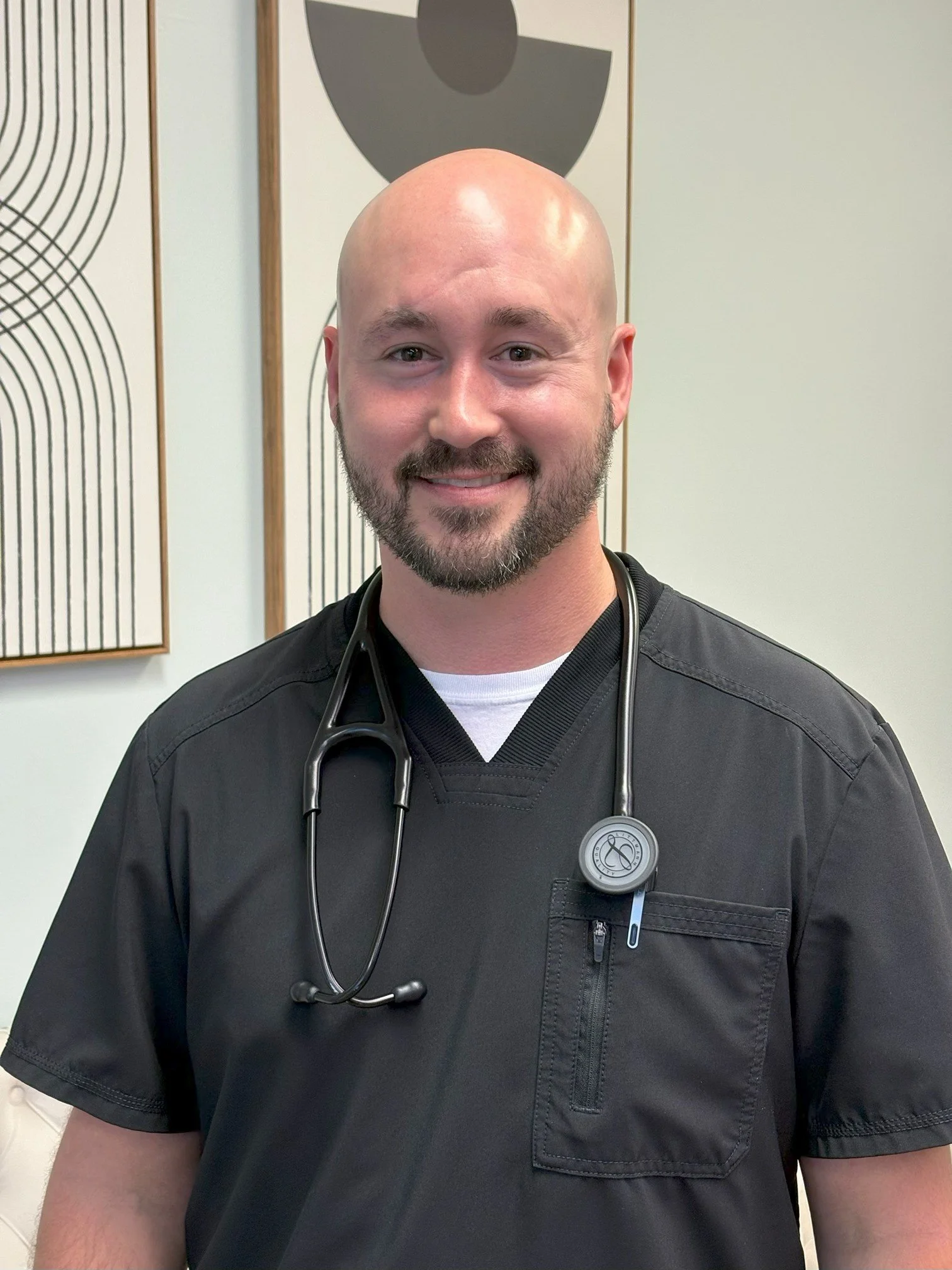 A male healthcare professional with a bald head and beard, wearing black scrubs and a stethoscope around his neck, smiling in an indoor setting with abstract wall art in the background.