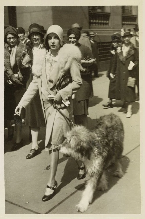 Black and white photograph of a woman in 1920s dress holding a cigarette and walking a large dog during the 1929 New York City Easter Parade, surrounded by a crowd of onlookers