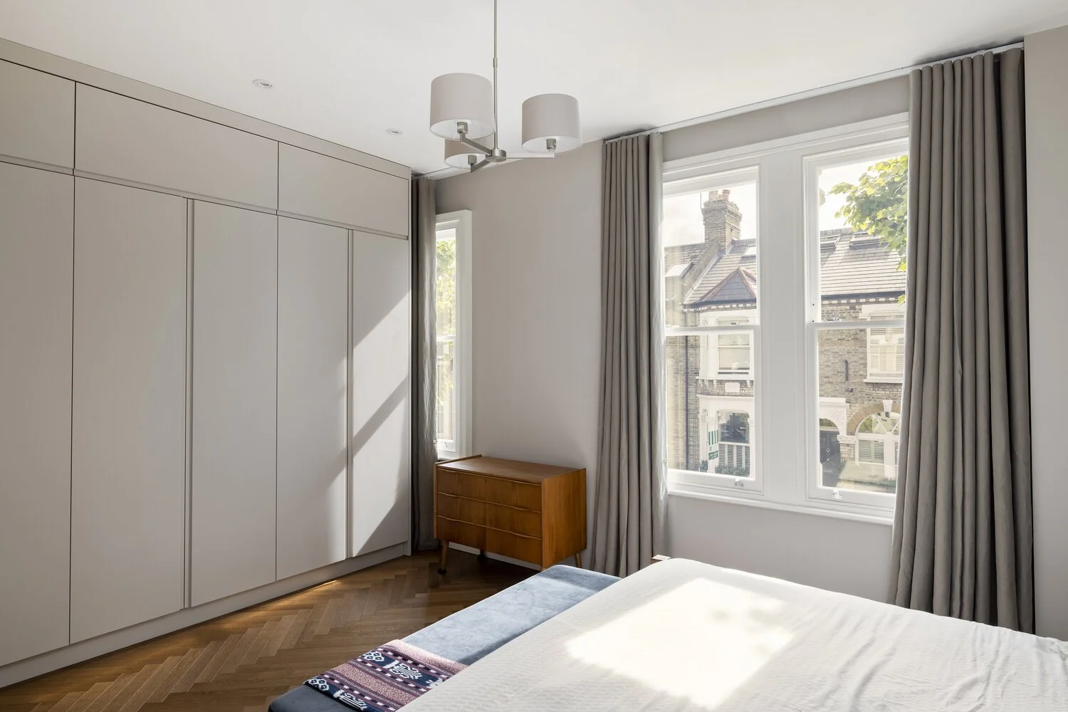  Bedroom  Bright bedroom with large windows, white wardrobe, wooden dresser, and bed with a colorful rug at the foot of the bed, in a modern home.