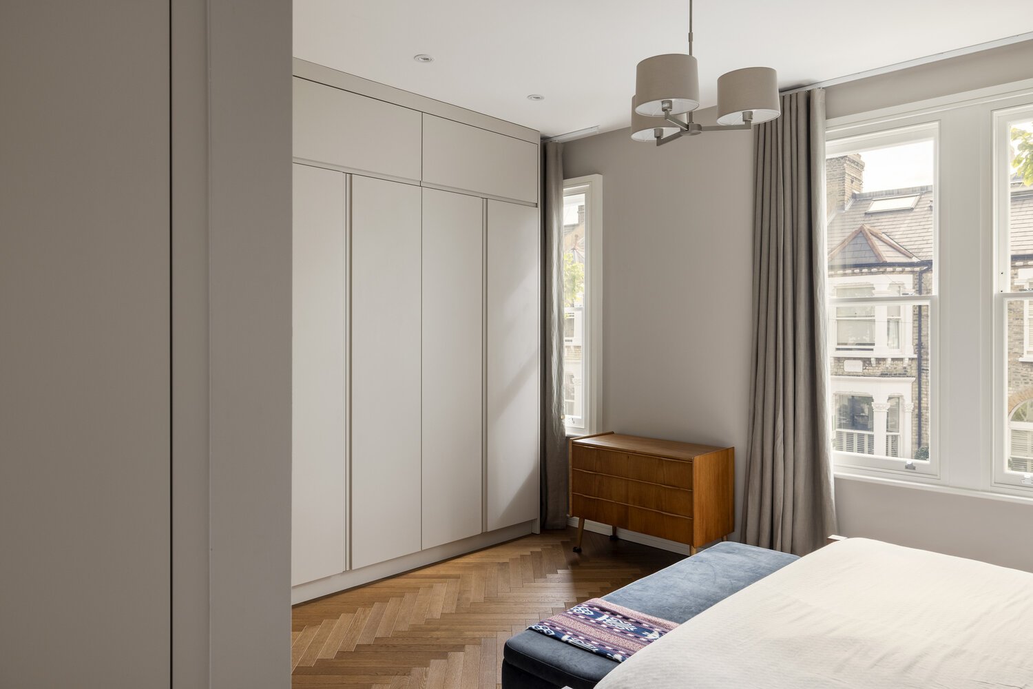  Bedroom  A bedroom with a white wardrobe, a wooden dresser, a bed with bedding, large windows with curtains, and a ceiling light fixture.