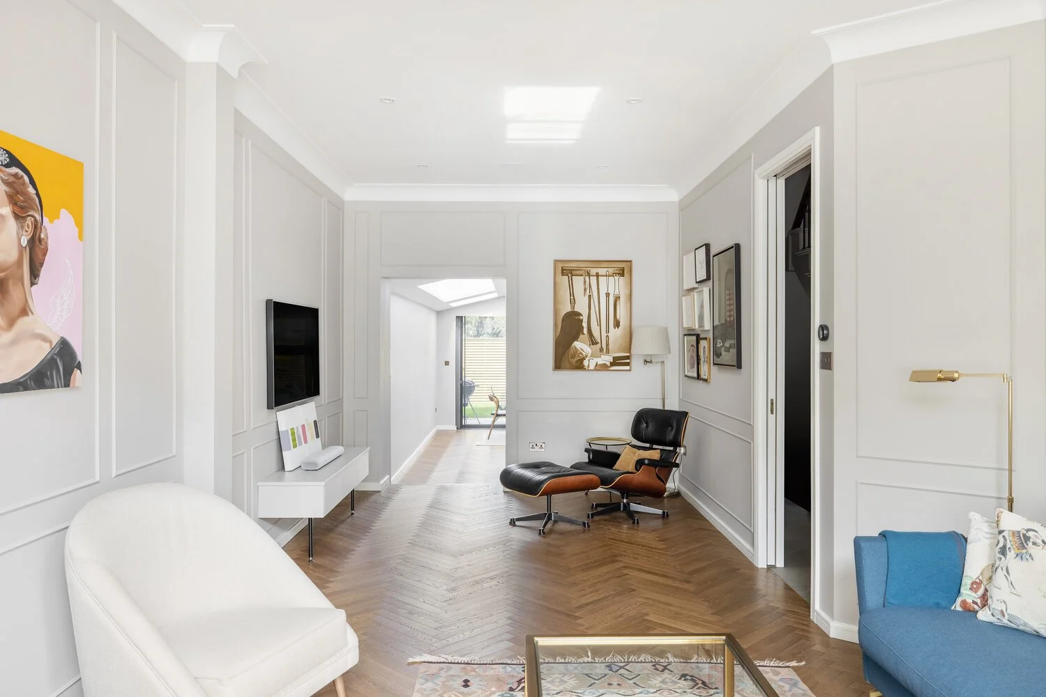  Living Room Living room with white walls, hardwood floor, wall-mounted TV, colorful artwork, black and tan lounge chair, blue sofa with floral pillows, white armchair, glass coffee table, and decorative wall art.