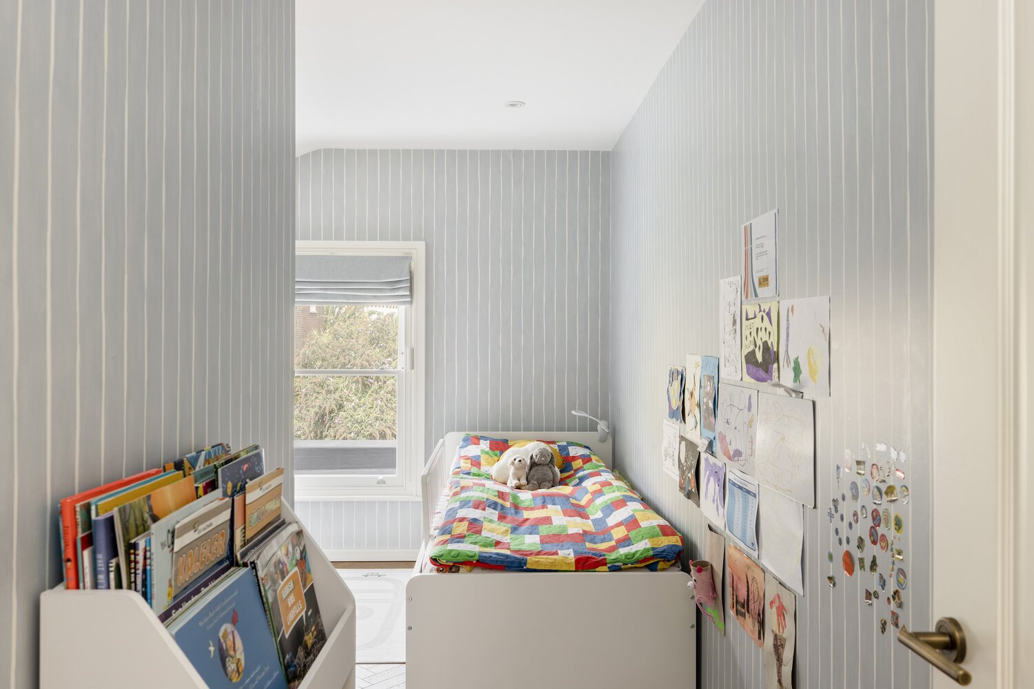  Bedroom  Child's bedroom with a white bed, colorful quilt, and plush toys, art on the walls, books on a nearby shelf, and a window with a gray blind showing trees outside.