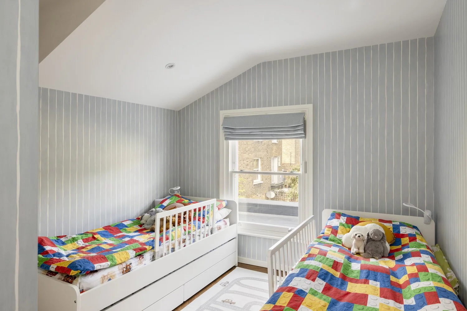  Bedroom  A bedroom with two twin beds with colorful Lego-themed bedding, plush toys on both beds, gray striped wallpaper, a window with a gray Roman shade, and a light-colored rug on a wooden floor.