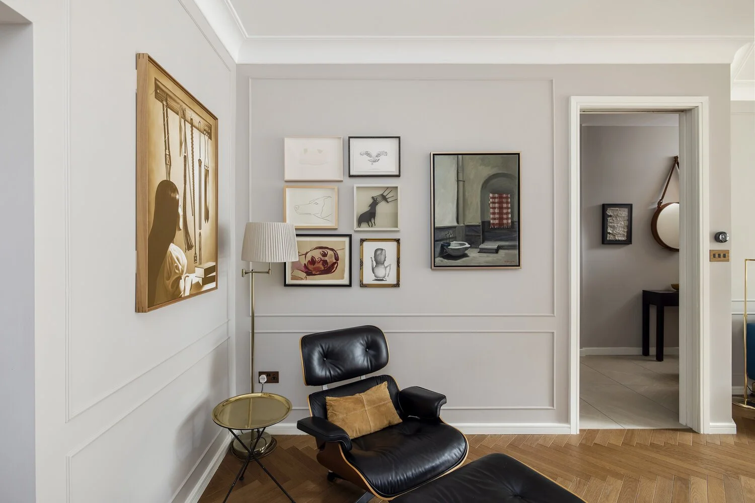  Living Room   Living room with white walls, framed artwork, a black leather lounge chair with a brown pillow, a small gold side table, a floor lamp, and an open doorway leading to another room with decor visible.