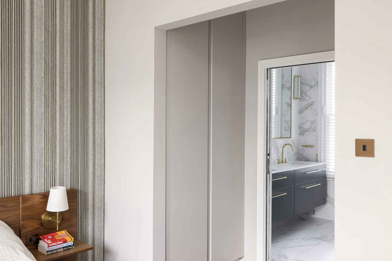  Bedroom  Bedroom with wooden headboard, small nightstand with books and a white lamp, textured wallpaper, door leading to a bathroom with marble walls and gold fixtures.