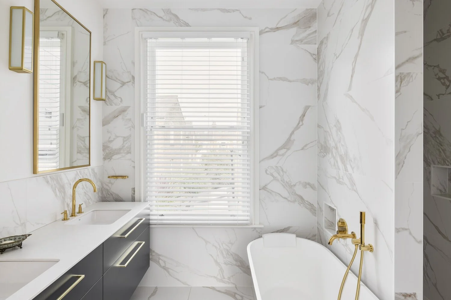  Bathroom Modern bathroom with white marble walls, a large window with white blinds, a white bathtub with gold fixtures, and a dark gray vanity with gold handles and a white countertop.
