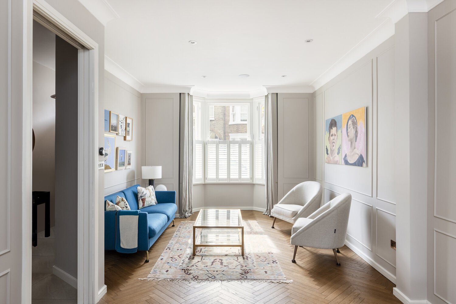  Living Room  Bright living room with white walls and hardwood floor, featuring a blue sofa, two white armchairs, a glass coffee table, a patterned rug, wall art, and a large bay window with shutters and curtains.