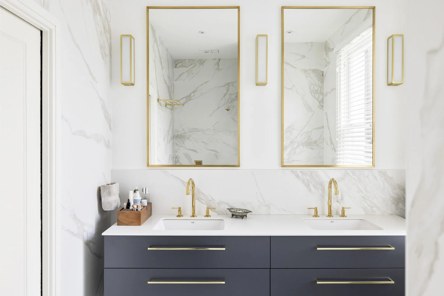  Bathroom Modern bathroom with twin sinks, gold faucets, and navy blue vanity cabinets. Large wall mirrors with gold frames, marble walls, and a window with blinds.