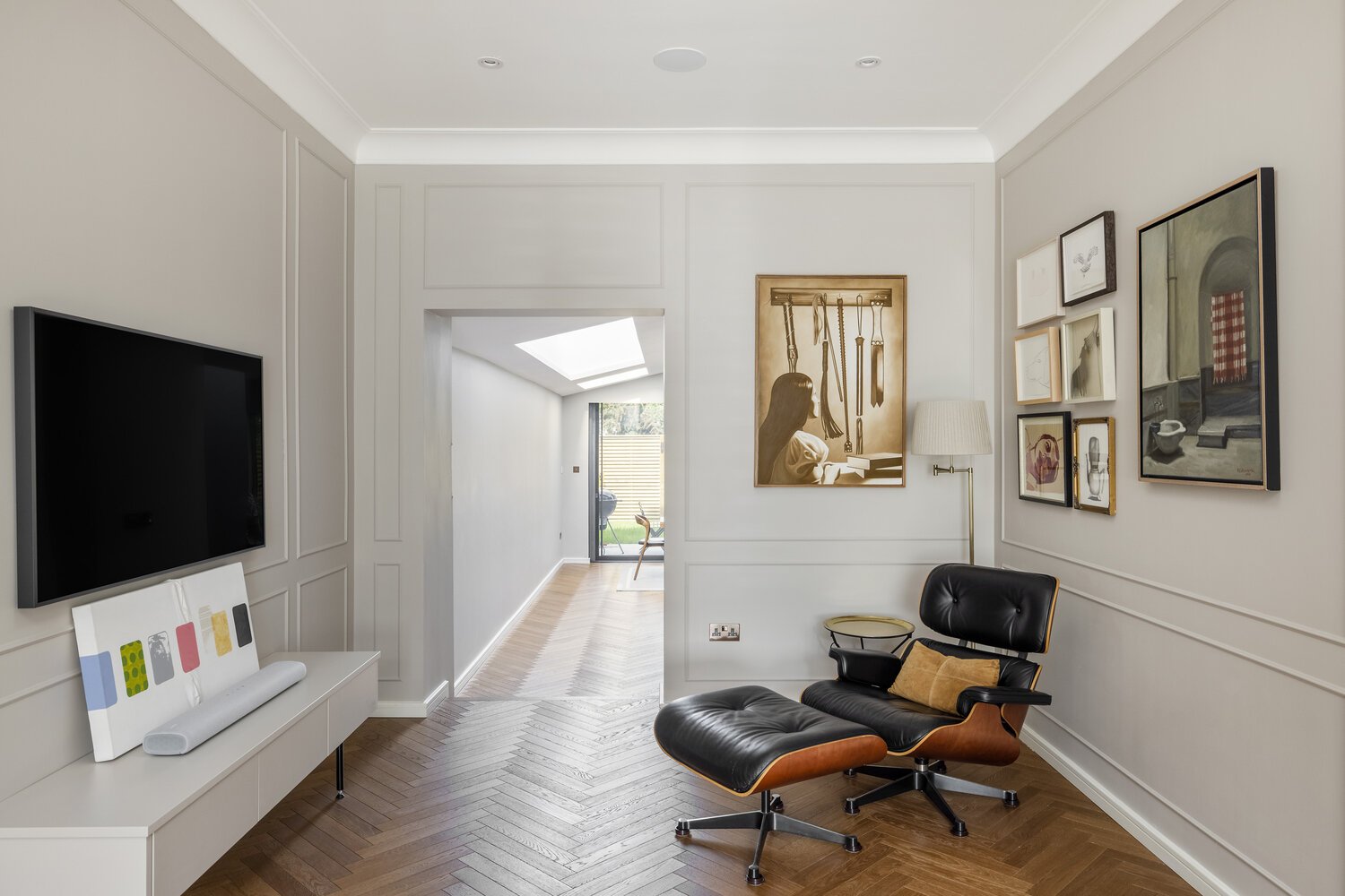  Living Room Living room with a black leather lounge chair, a white console with colorful art and a sound bar, a wall-mounted flat-screen TV, a floor lamp, and framed artwork on the wall. A hallway leading to a sunlit space with a table and chairs is