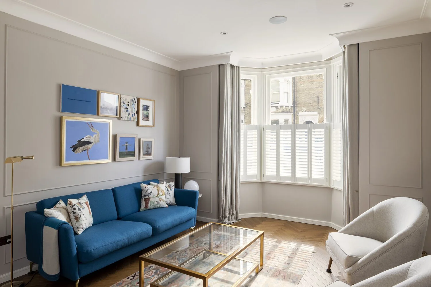  Living Room  Living room with blue sofa, white armchair, glass coffee table, wall art, large window with white shutters, and light-colored curtains.