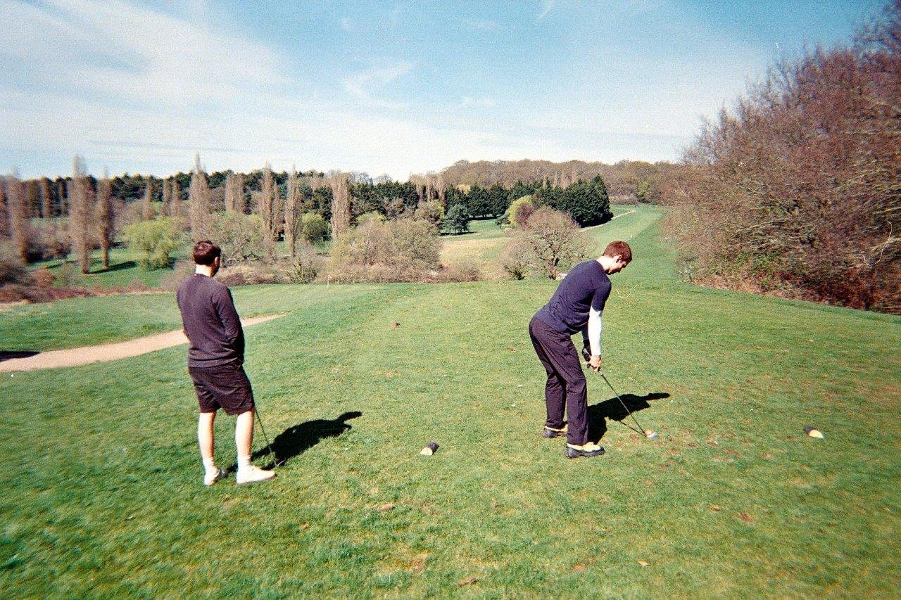 Two male golfers standing at a par 3 tee box, one is ready to hit the ball, the other is looking down towards the green in anticipation.